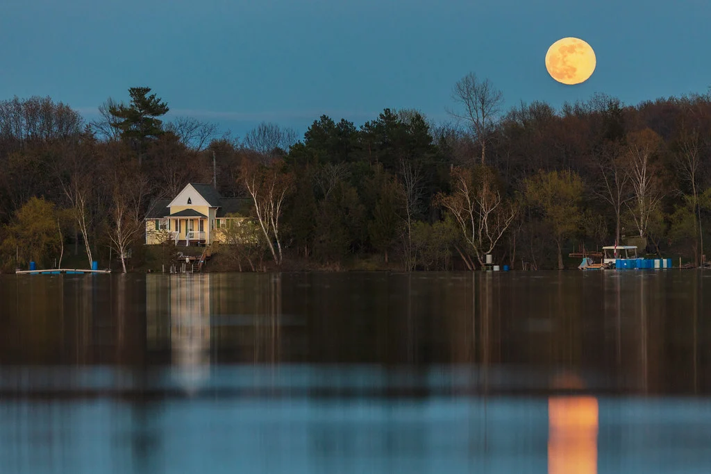 A full moon rising over a lakeside cottage at dusk, reflected in the calm water