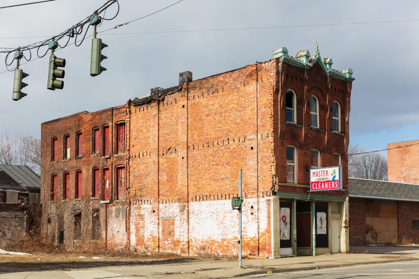 A three-story red brick commercial building with boarded and empty windows, patches of faded white paint along the base and a dry cleaner's sign on the corner, with traffic lights hanging in the foreground