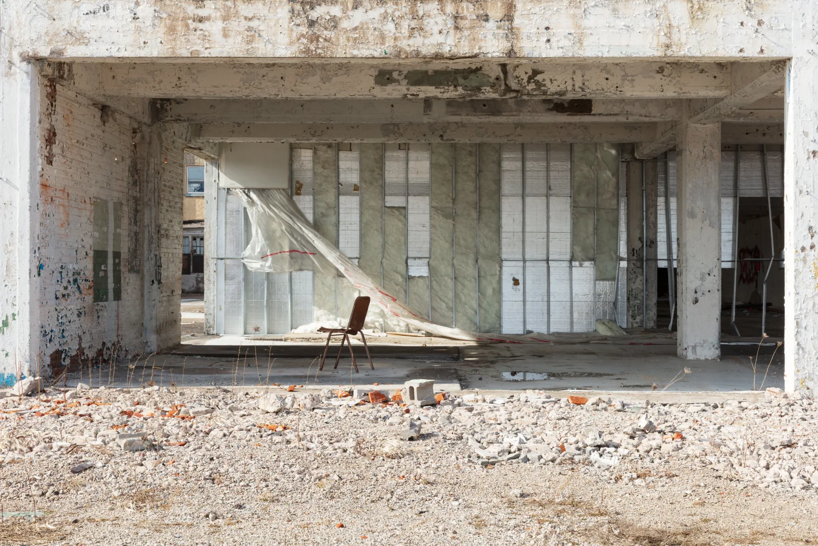 The open ground floor of a gutted concrete building with a single folding chair standing on the bare slab, torn plastic sheeting hanging behind it and a field of rubble and broken bricks in the foreground