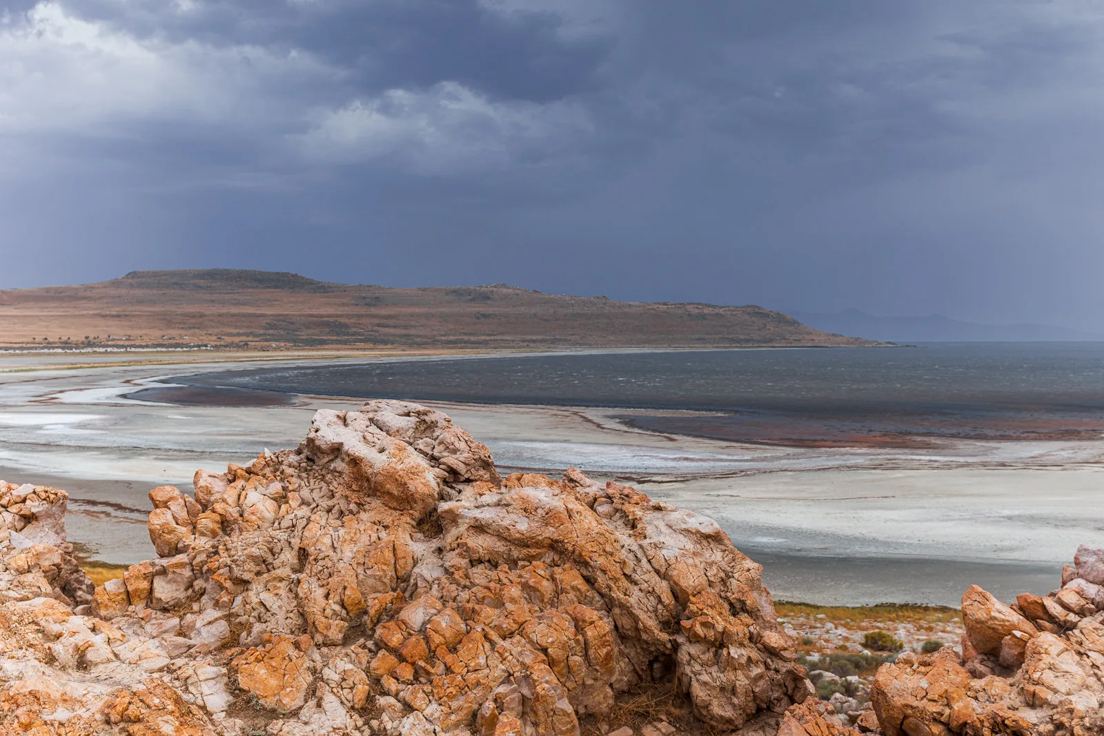 Orange, craggy rocks in the foreground, a pale shoreline and grey lake in the middle, and a brown hill under dark storm clouds behind