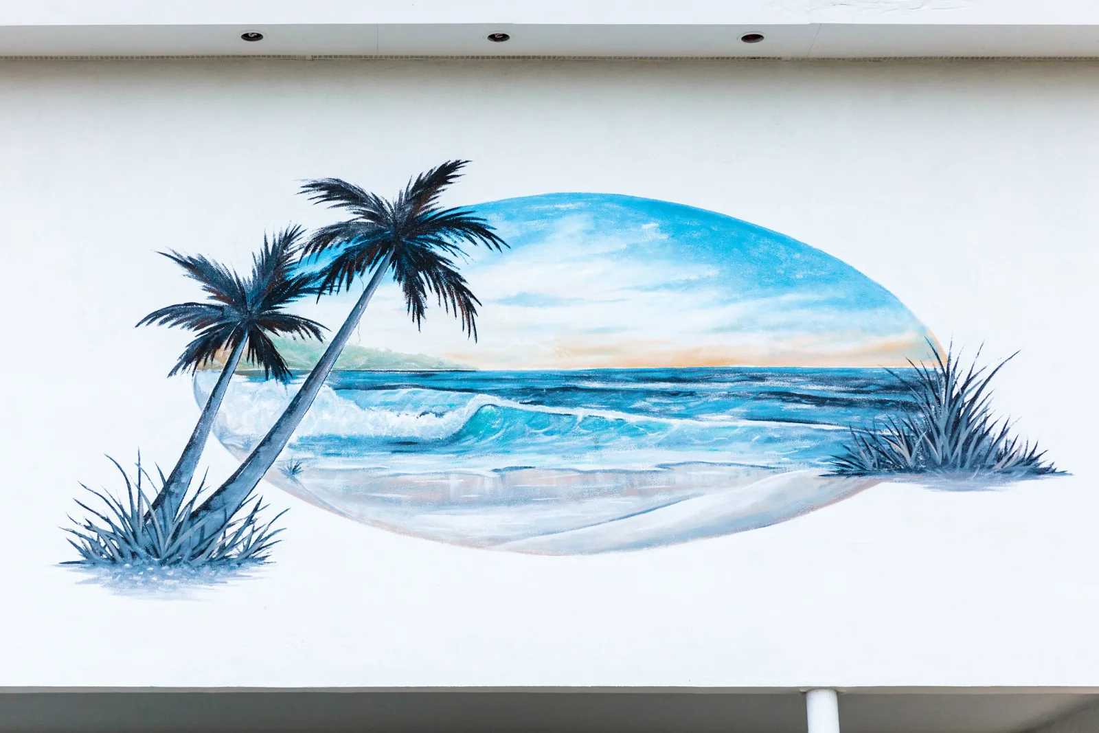An oval painting of a beach with palm trees and breaking waves on a plain white wall below a flat roofline