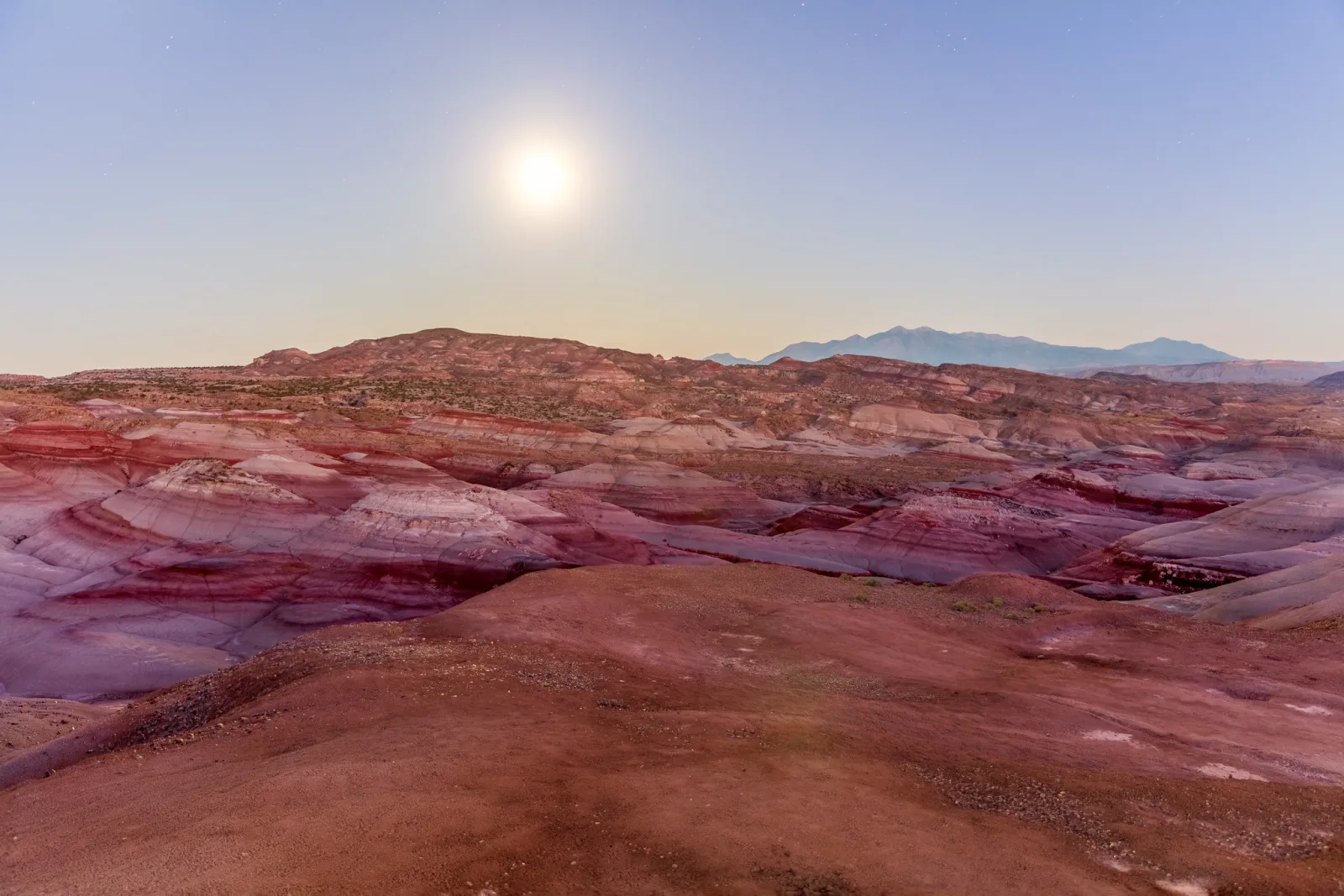 Striped red, purple and grey clay hills stretch toward distant mountains under a bright moon in a pale blue sky, the landscape looking almost like daylight