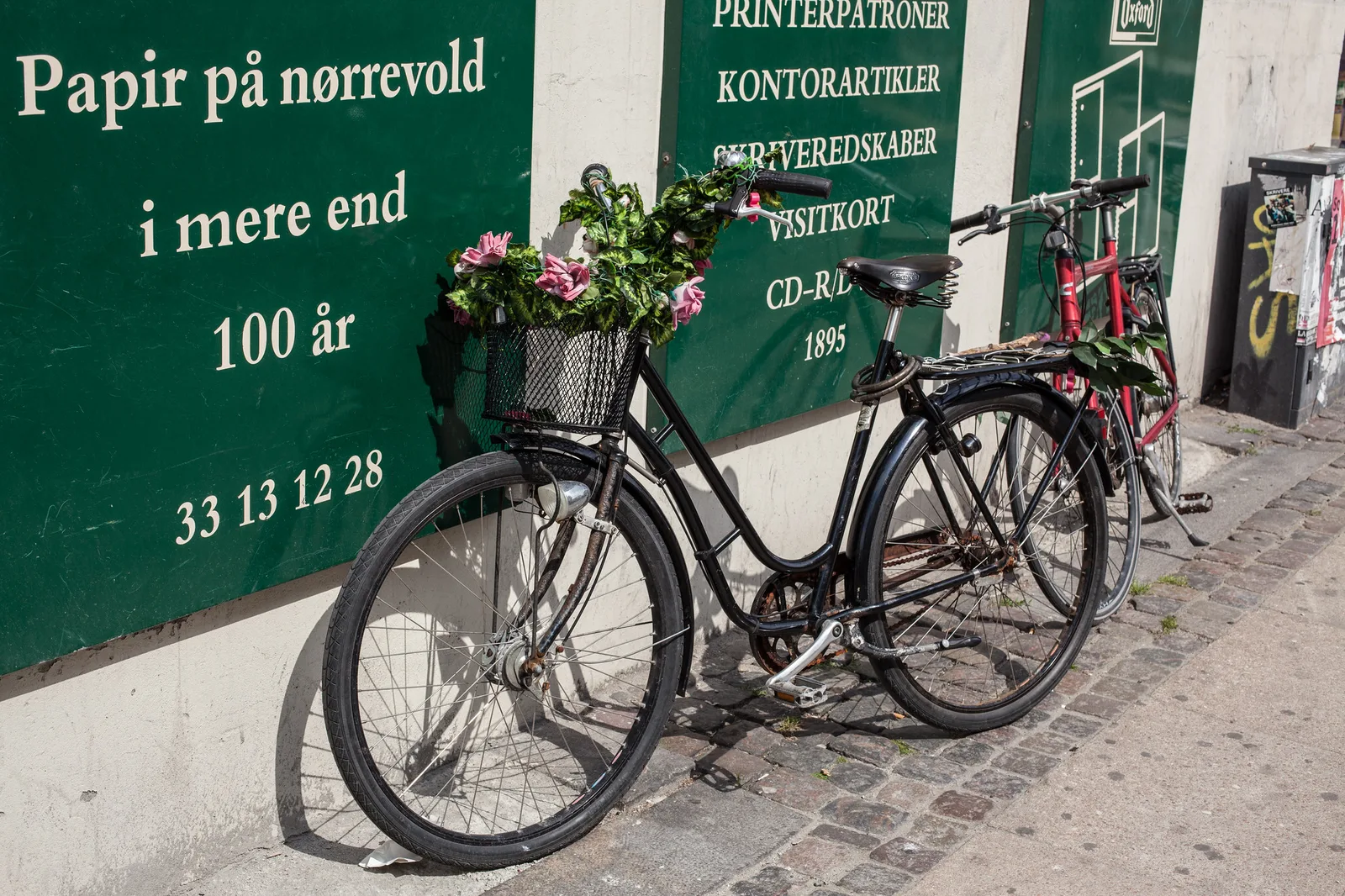 A black bicycle with a basket of pink flowers leaning against a wall with a green painted shop sign, on a cobbled pavement