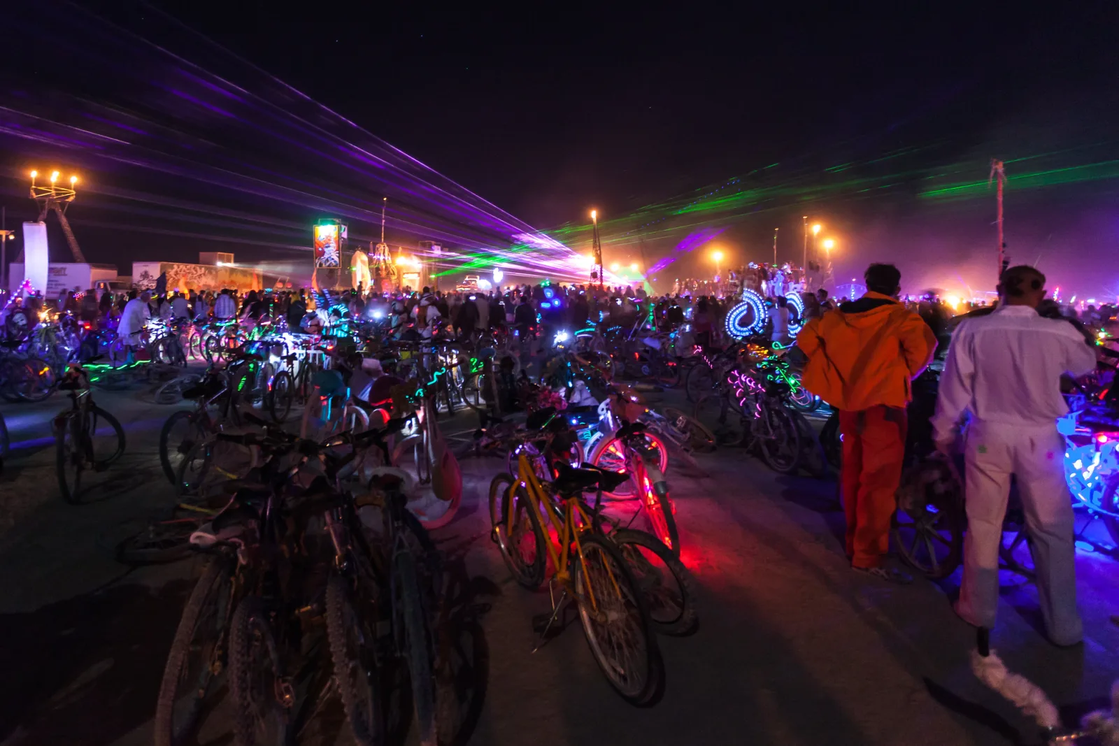 At night, rows of parked bicycles fill the foreground, two people in orange and white suits stand in the middle distance, and a crowd under purple and green laser beams fills the background