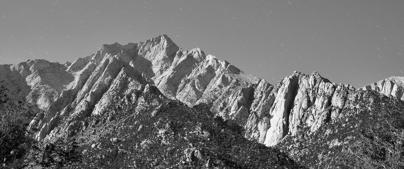 A black and white panorama of a jagged granite mountain with sharply lit ridges and shaded gullies above dark scrub-covered slopes