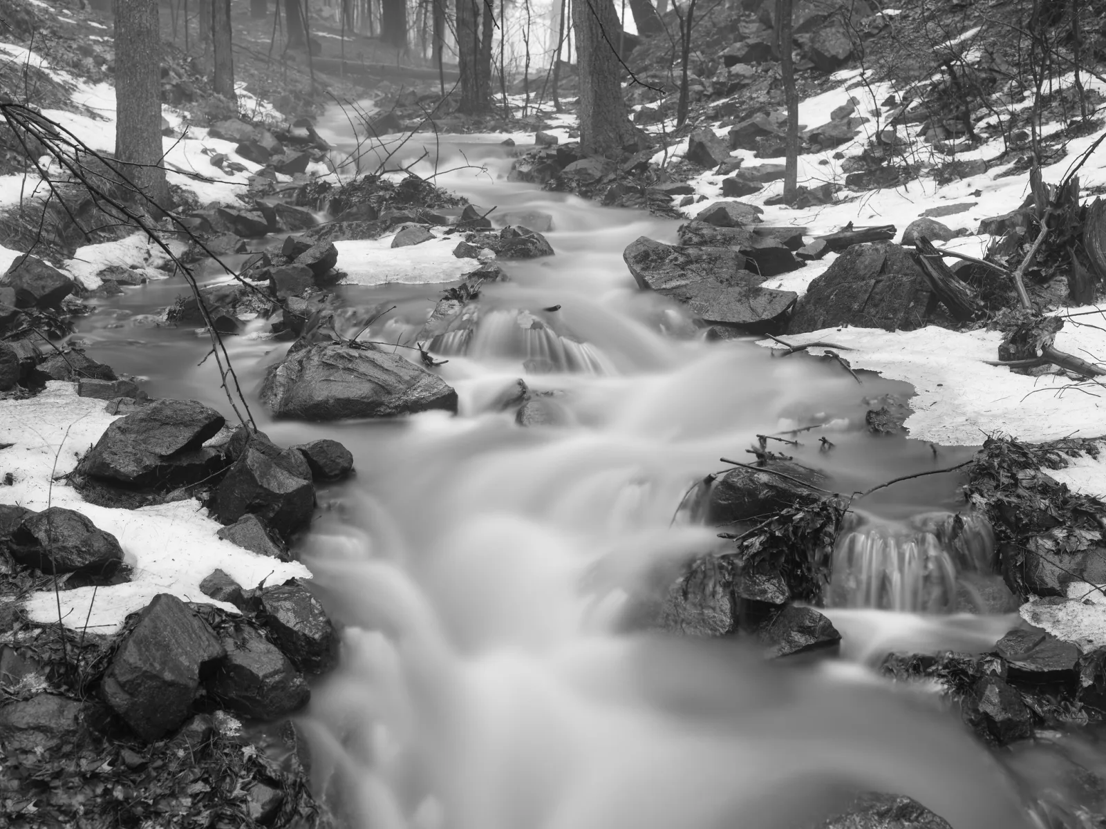 Black and white photo of a stream flowing as smooth white water between dark rocks, patches of snow and bare tree trunks