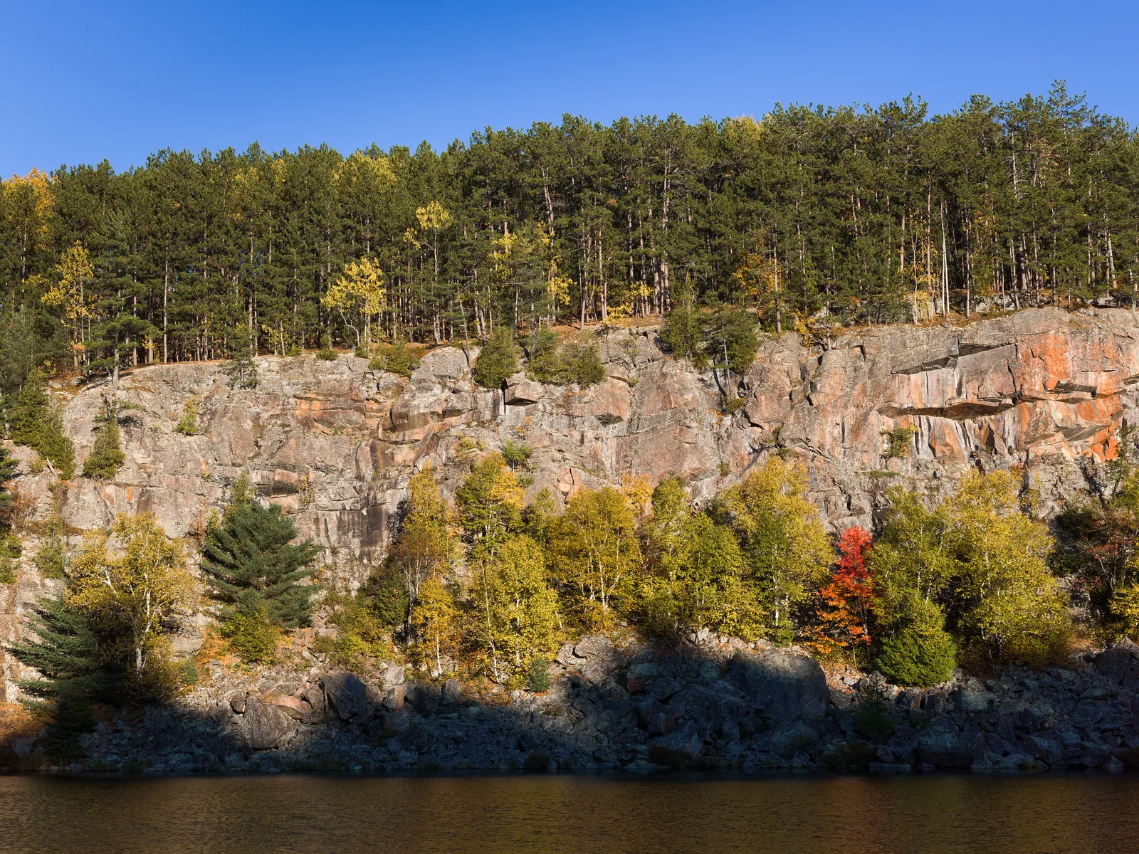 A deep blue sky above a ridge of pine trees on a sunlit orange rock cliff, with autumn trees and dark water below