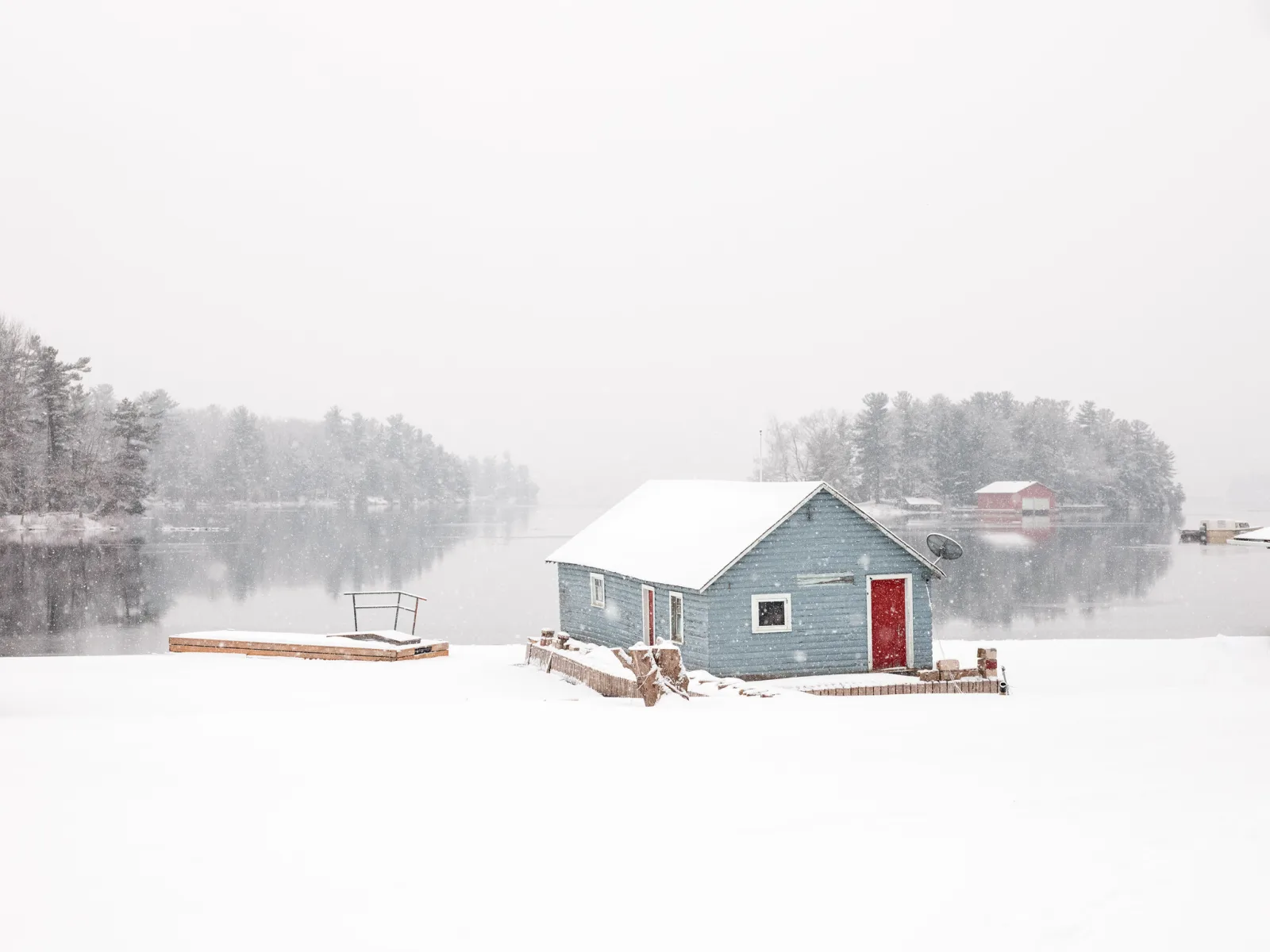 A small blue-grey boathouse with a red door on a snowy shore, pale misty water and faint snowy islands behind