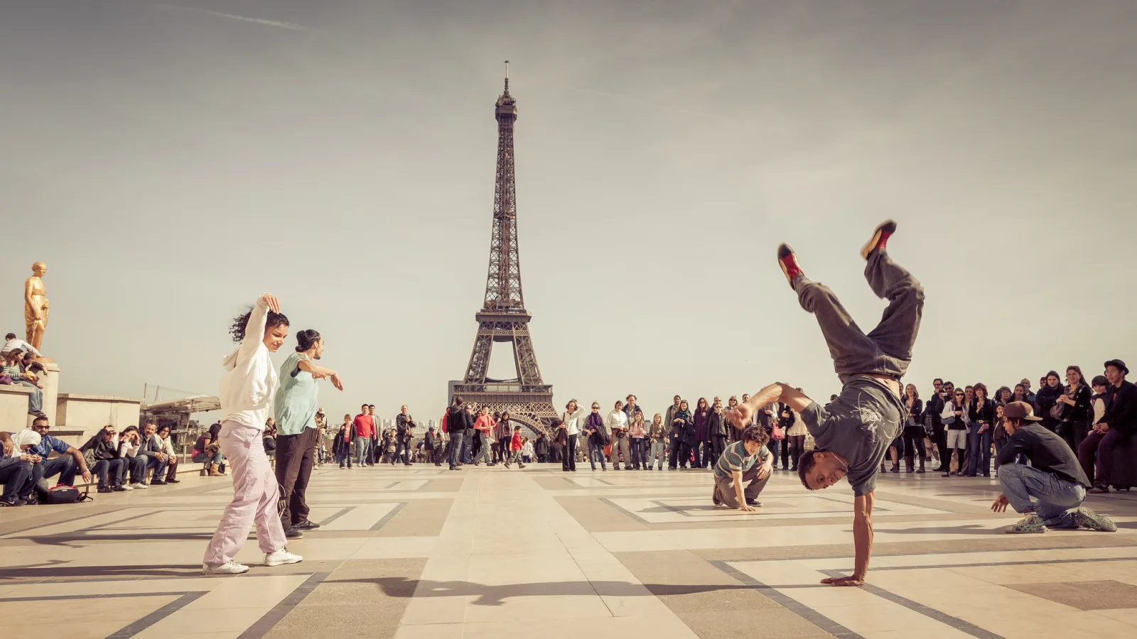 A breakdancer balanced on one hand with legs raised in the air on a plaza, a crowd watching and the Eiffel Tower in the background