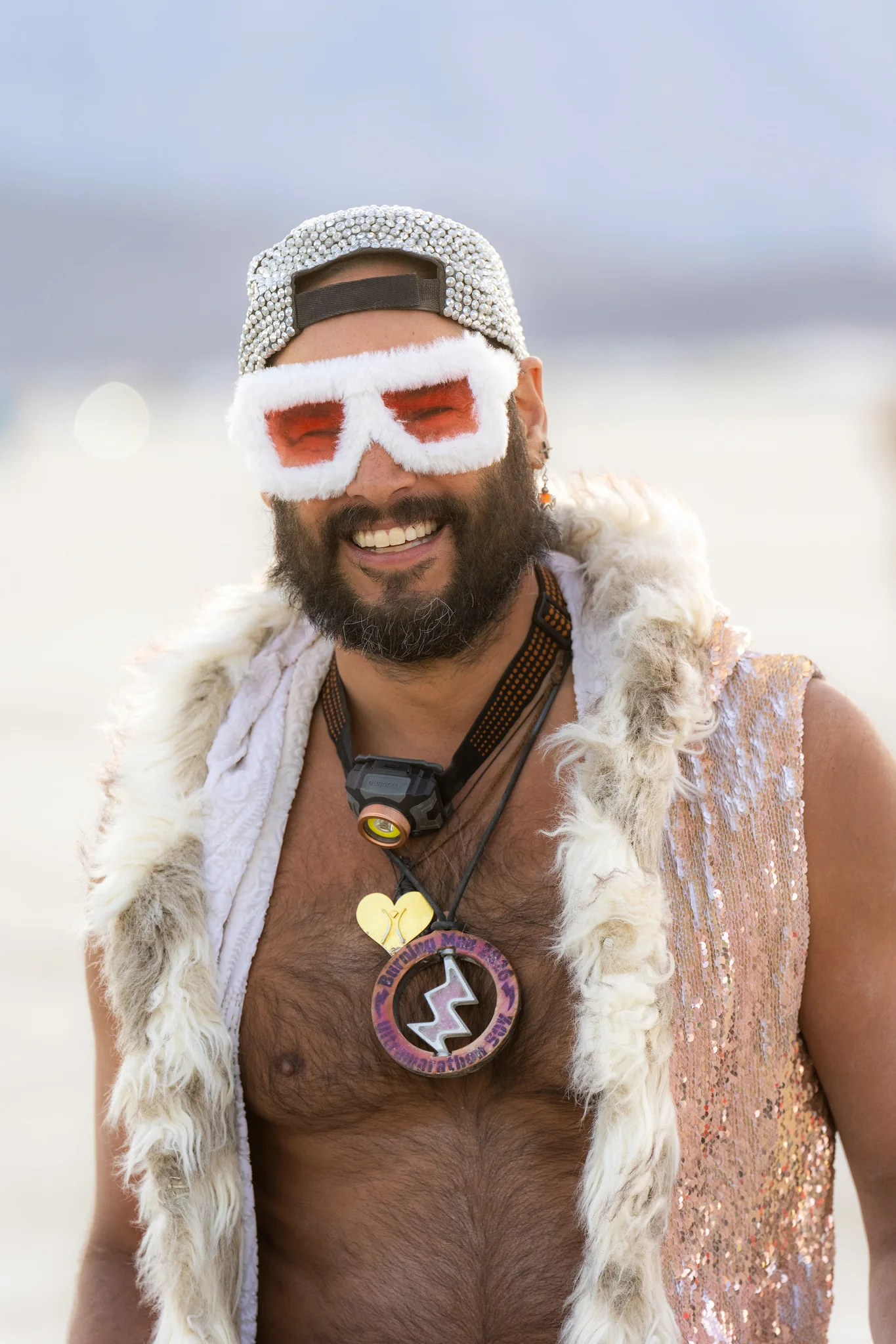 A smiling bearded man in white-framed novelty glasses and a fur vest stands in front of a very bright, pale desert background