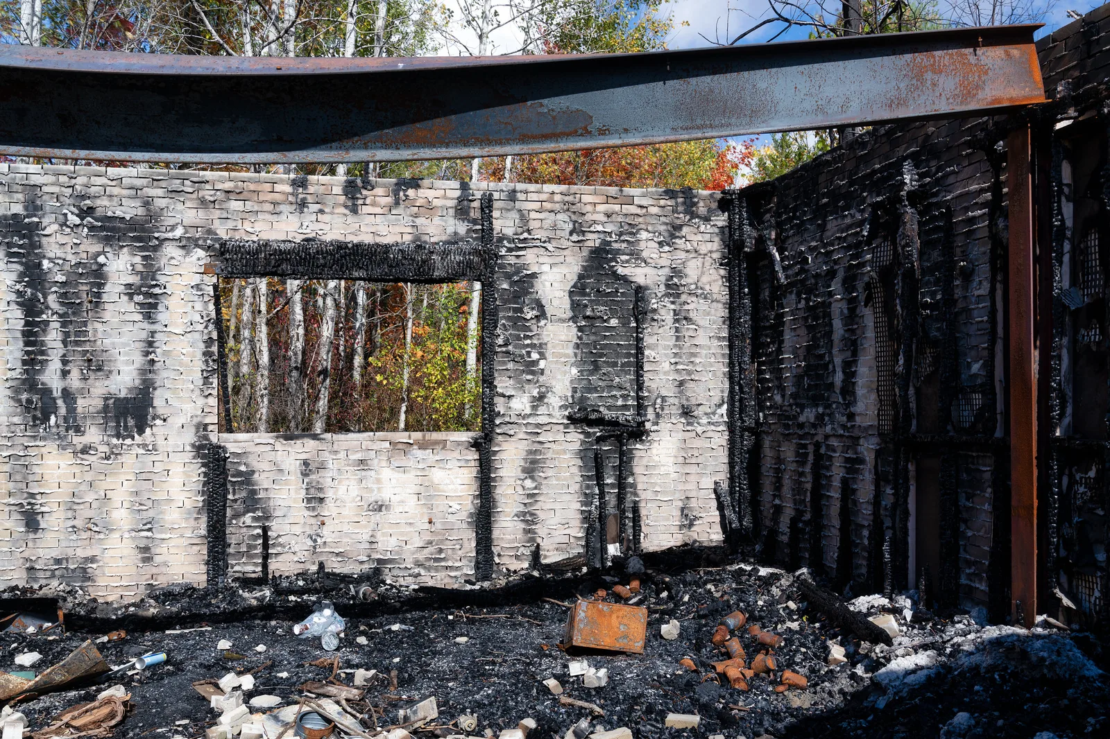 The roofless interior of a burnt-out brick building with scorched walls, an empty window opening, a rusted steel beam and charred debris on the floor