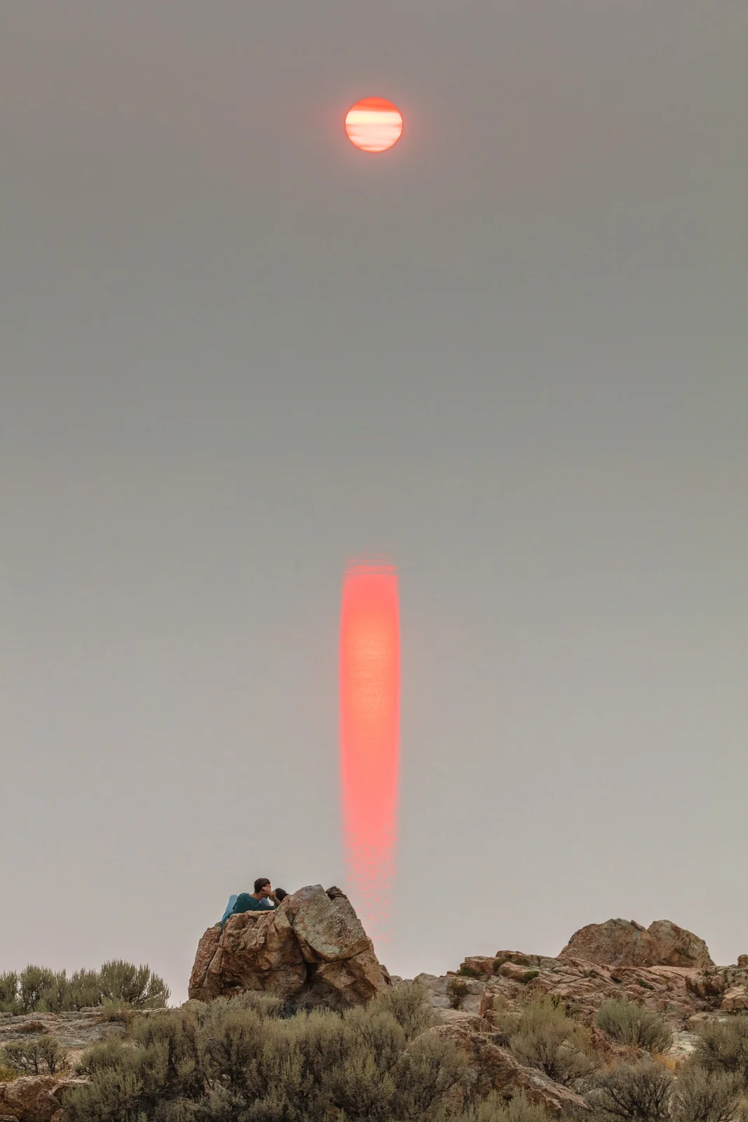 A silhouetted couple sitting on a rock formation with a bright sun and its reflection glowing above them in a hazy desert sky