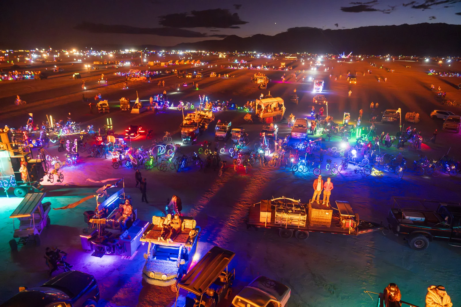 A wide nighttime desert scene lit by colorful vehicle and art lighting under a dark sky