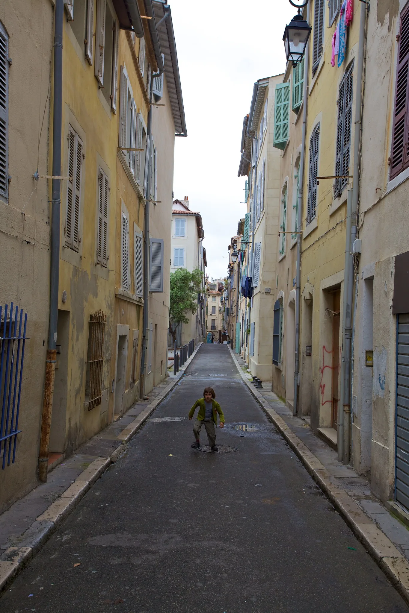 A narrow street between tall old buildings with a small child in the middle of the road, the walls and pavement converging behind the child