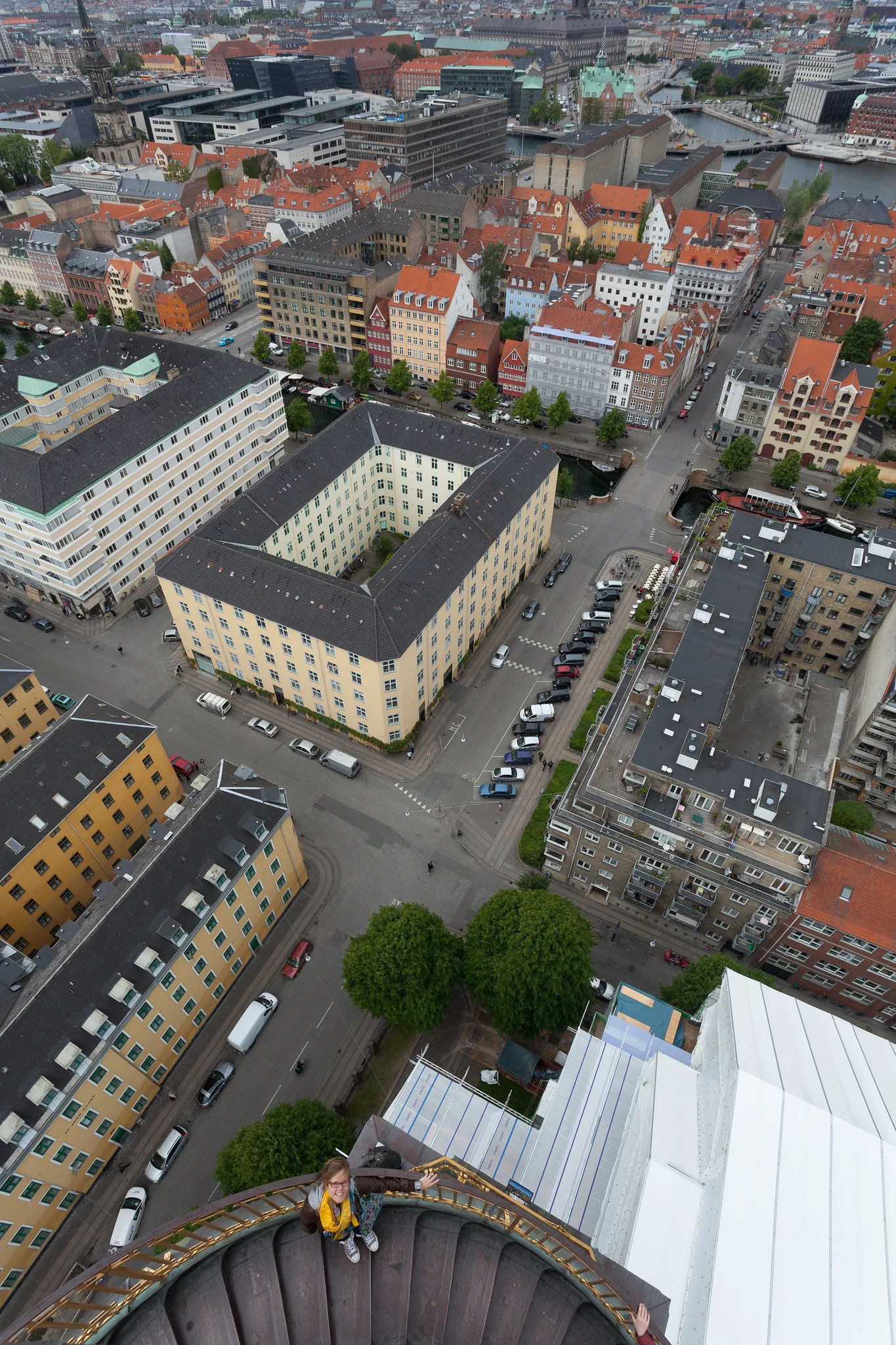 A view straight down over a dense city centre of red-tiled roofs, courtyard blocks and streets with small parked cars, taken from the top of a tower