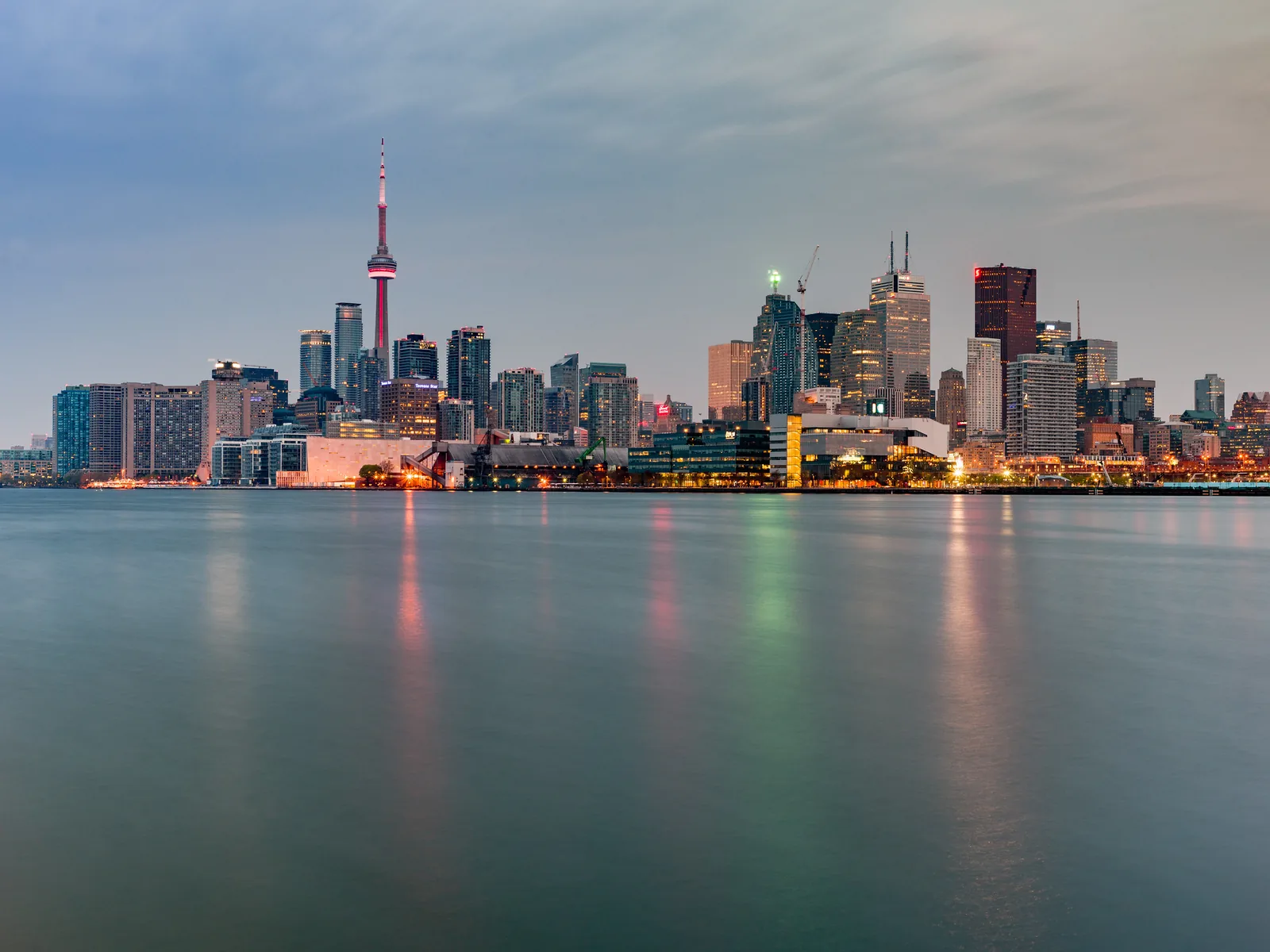A city skyline with a tall tower seen across calm water at dawn, building lights still on and reflected as coloured streaks in the smooth water under a grey-blue sky