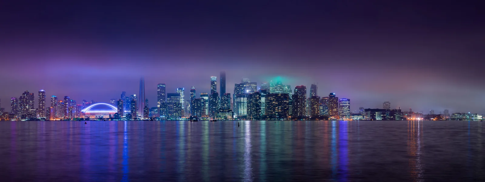 A city skyline at night seen across a wide stretch of water, with coloured reflections on the surface