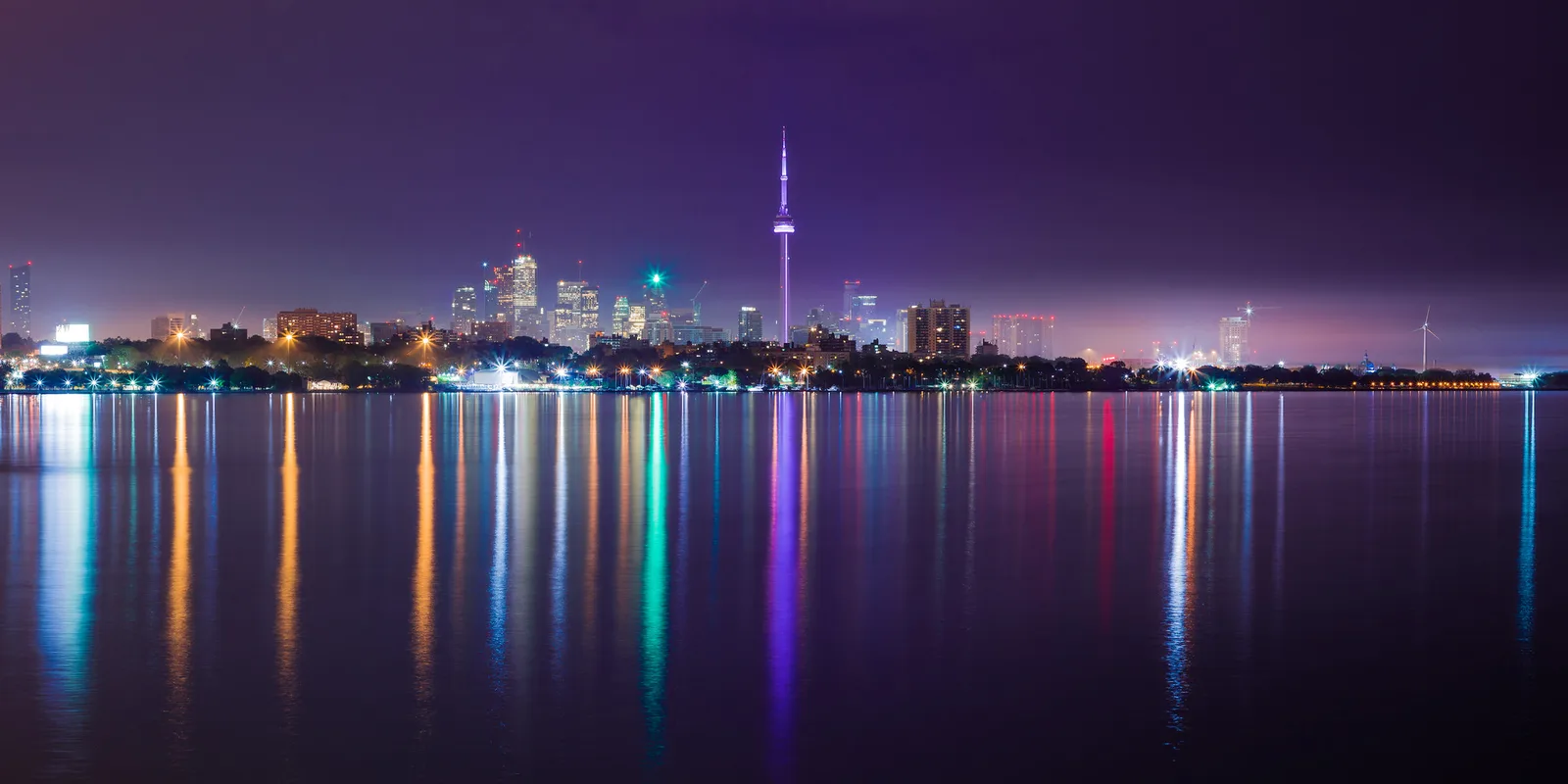 A city skyline at night across a wide stretch of dark water, with a tall tower in the centre and small bright lights strung along the whole shoreline, reflected as coloured streaks