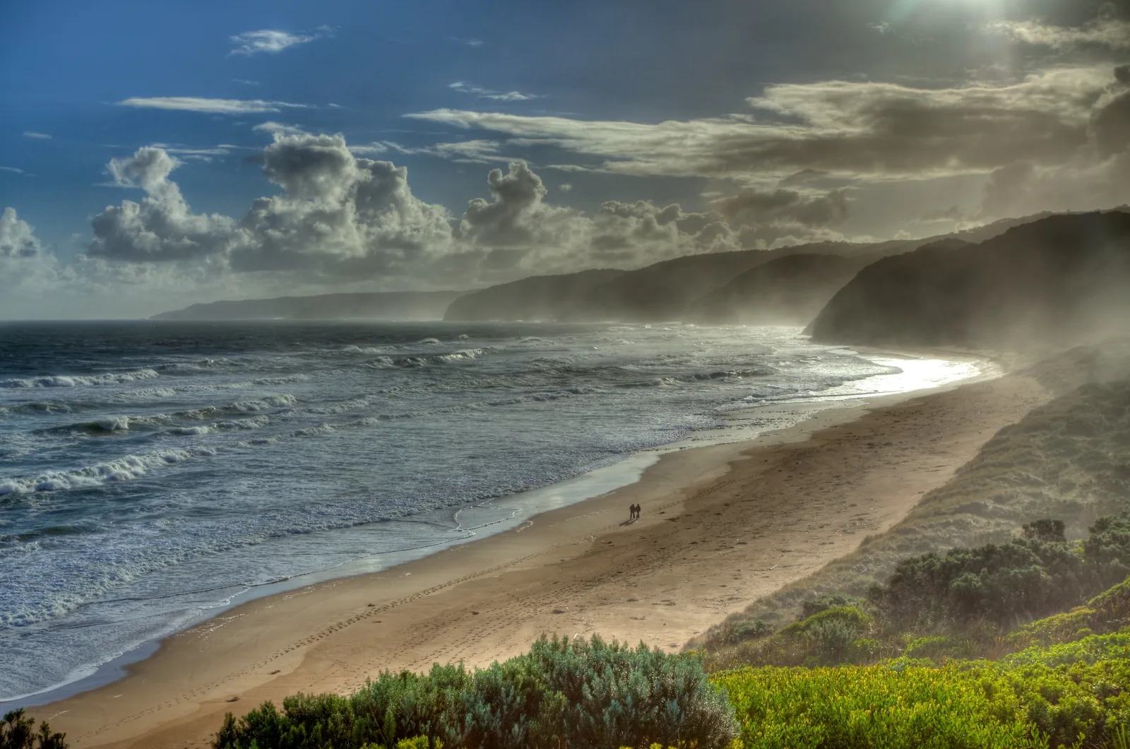 A long curving beach with waves rolling in, sea mist and spray haze drifting over the headlands, dramatic clouds above