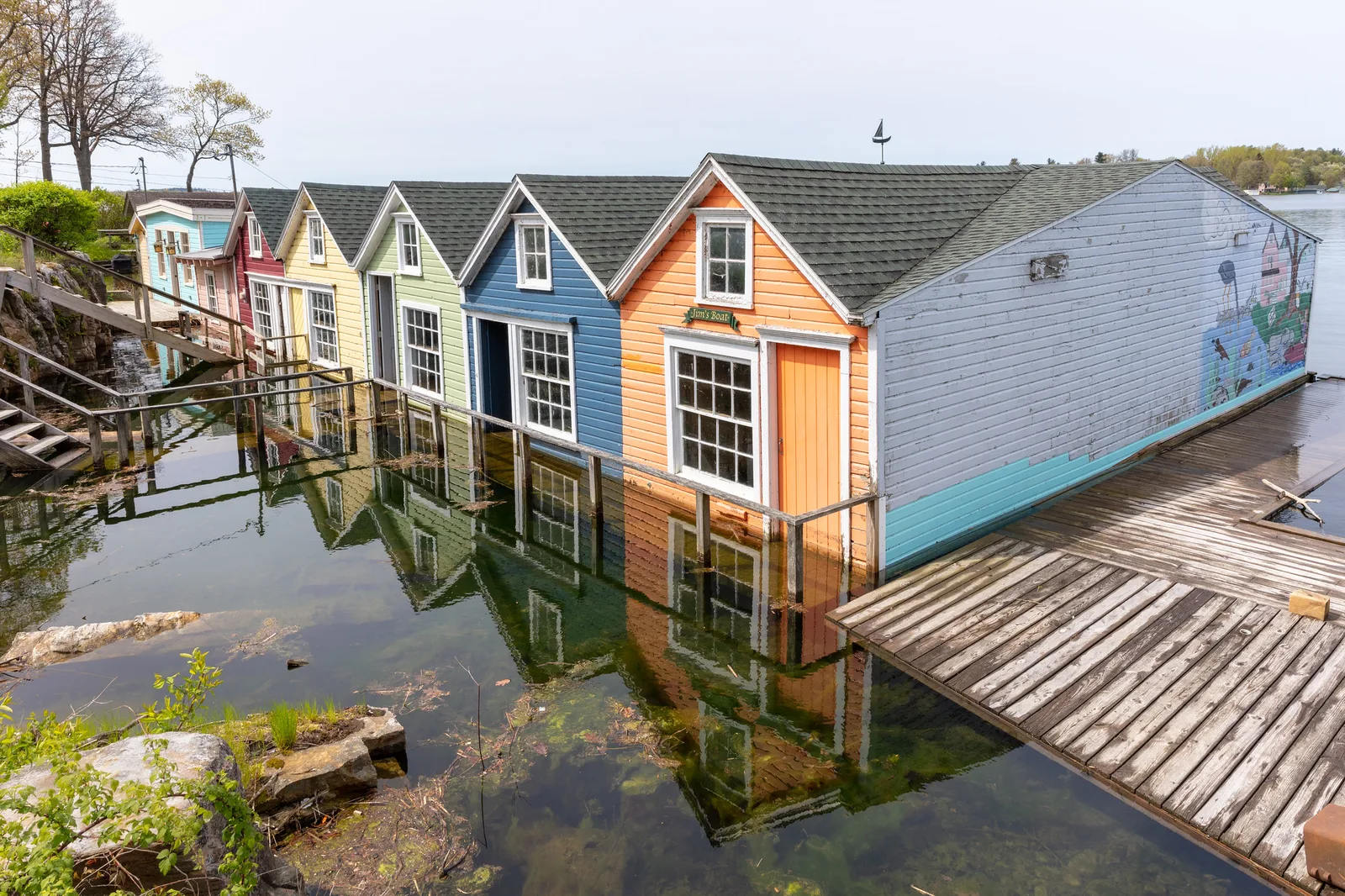 A row of wooden boathouses painted orange, blue, green, yellow and red standing in calm water that reflects them, beside a weathered wooden dock