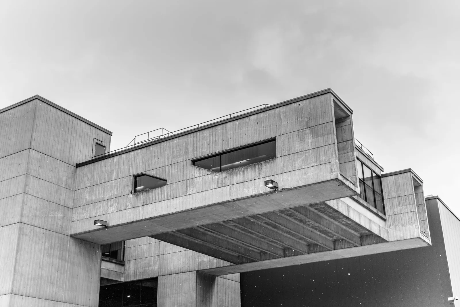 A black and white view looking up at a concrete building with stacked, overhanging box shapes against a pale cloudy sky