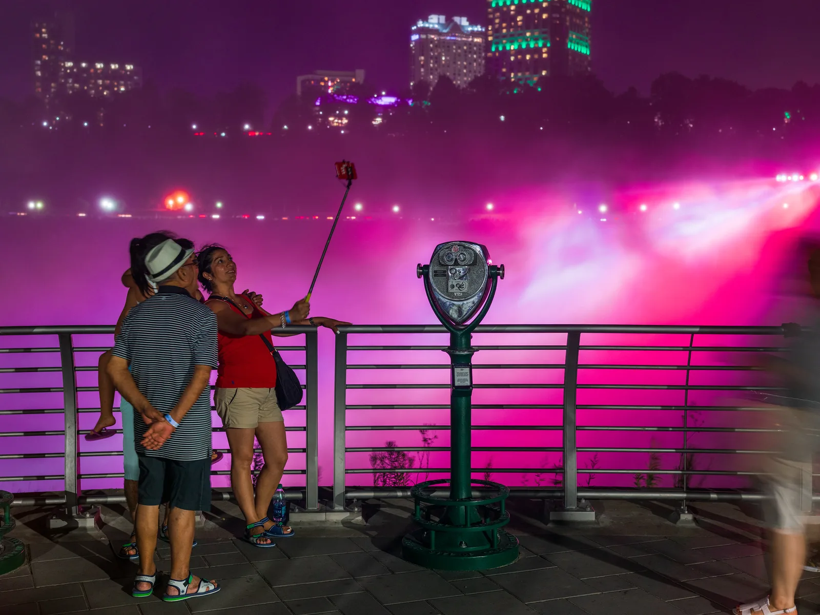 Two people at a railing at night raising a phone on a selfie stick in front of mist lit magenta, with a blurred passer-by at the edge of the frame