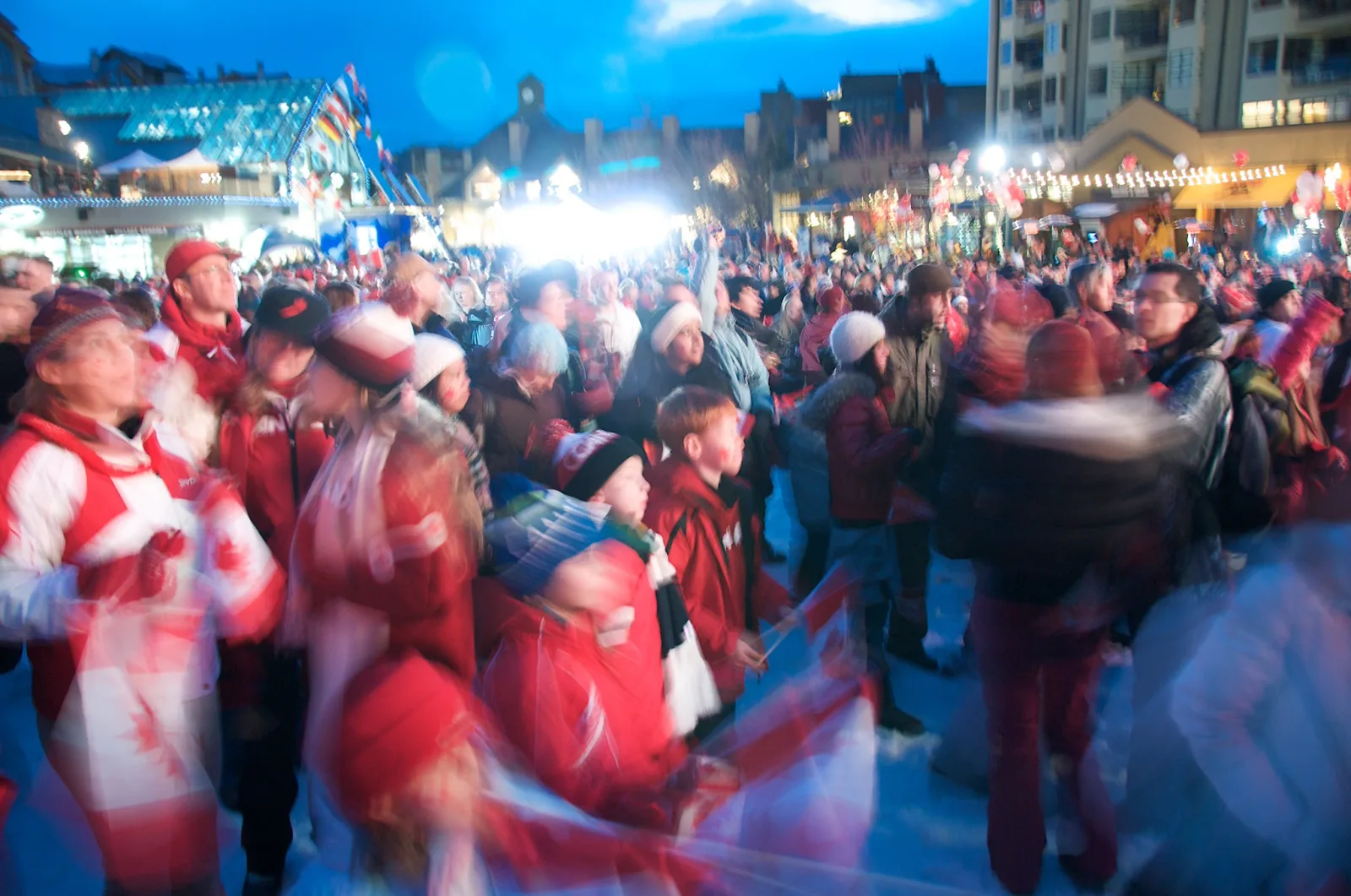 A dense crowd in red and white gathered in a lit city square at dusk, with many people blurred by movement and flags swinging