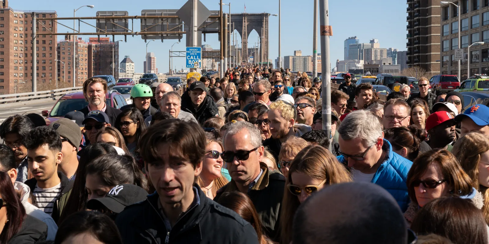 A dense crowd of people walking toward the camera on a bridge walkway, with the bridge's stone towers and city buildings behind