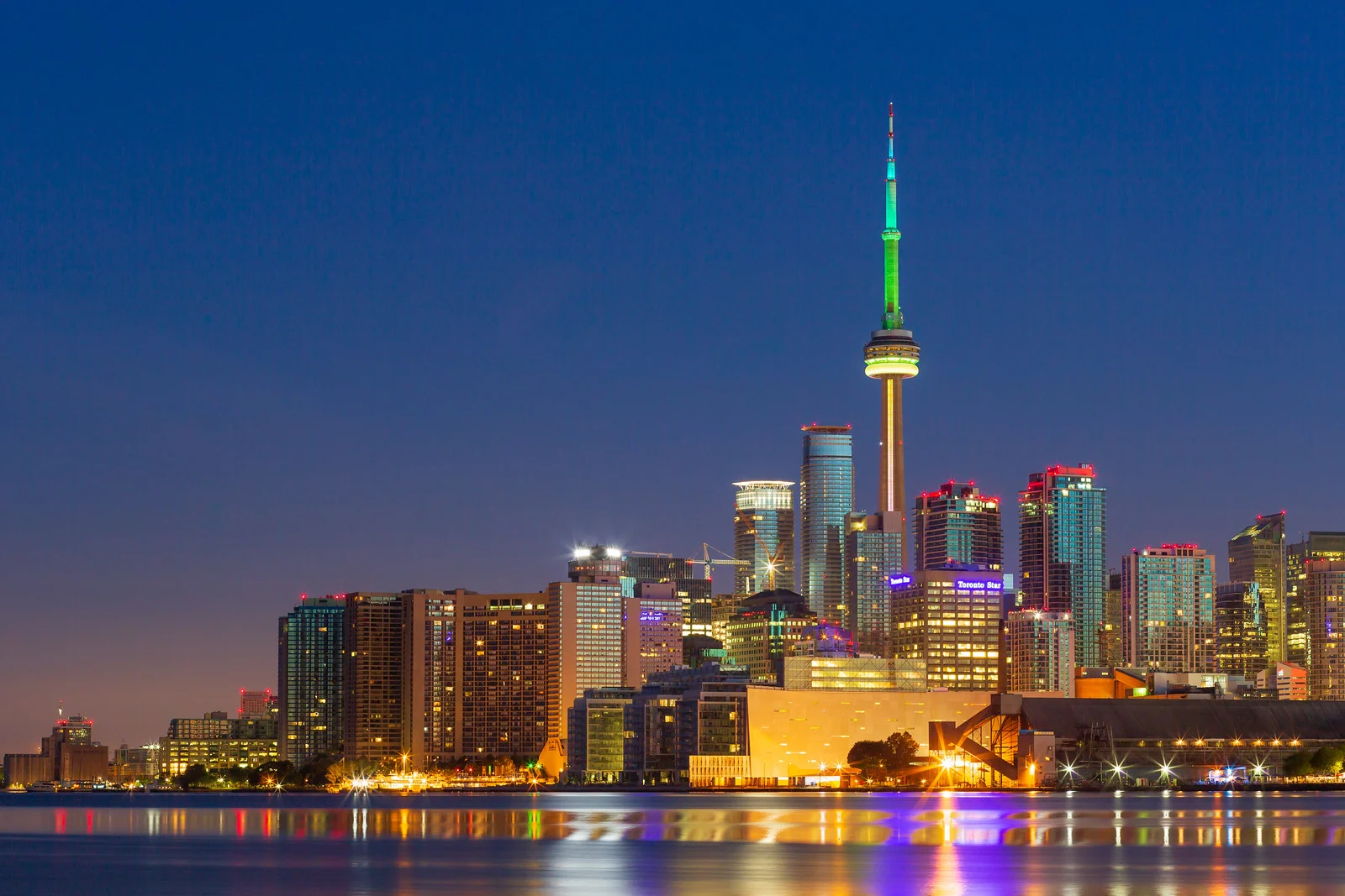 A city skyline at dawn with brightly lit towers and a glowing tower against a dark sky and dark water