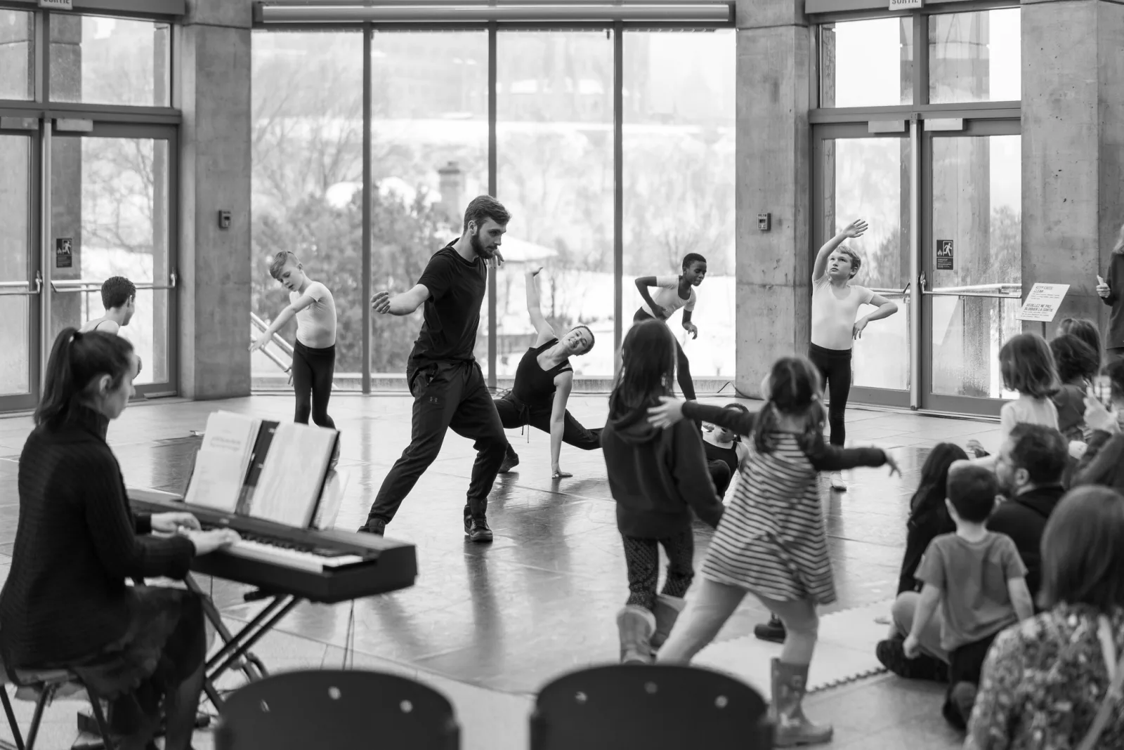 Black and white photo of a dance workshop in a bright glass-walled hall, a male instructor lunging forward as children copy the move and a pianist plays at left