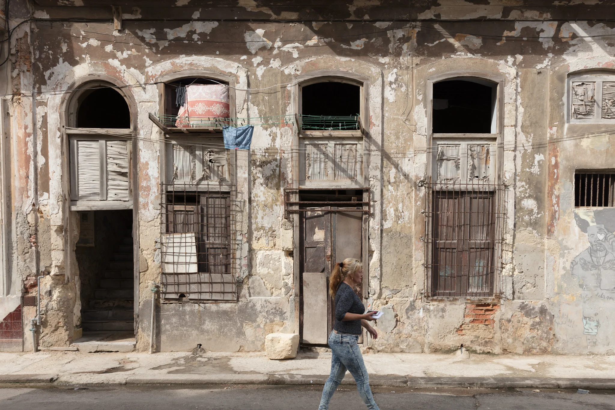 A woman walks along the sidewalk in front of a weathered, crumbling building facade with arched windows in Havana