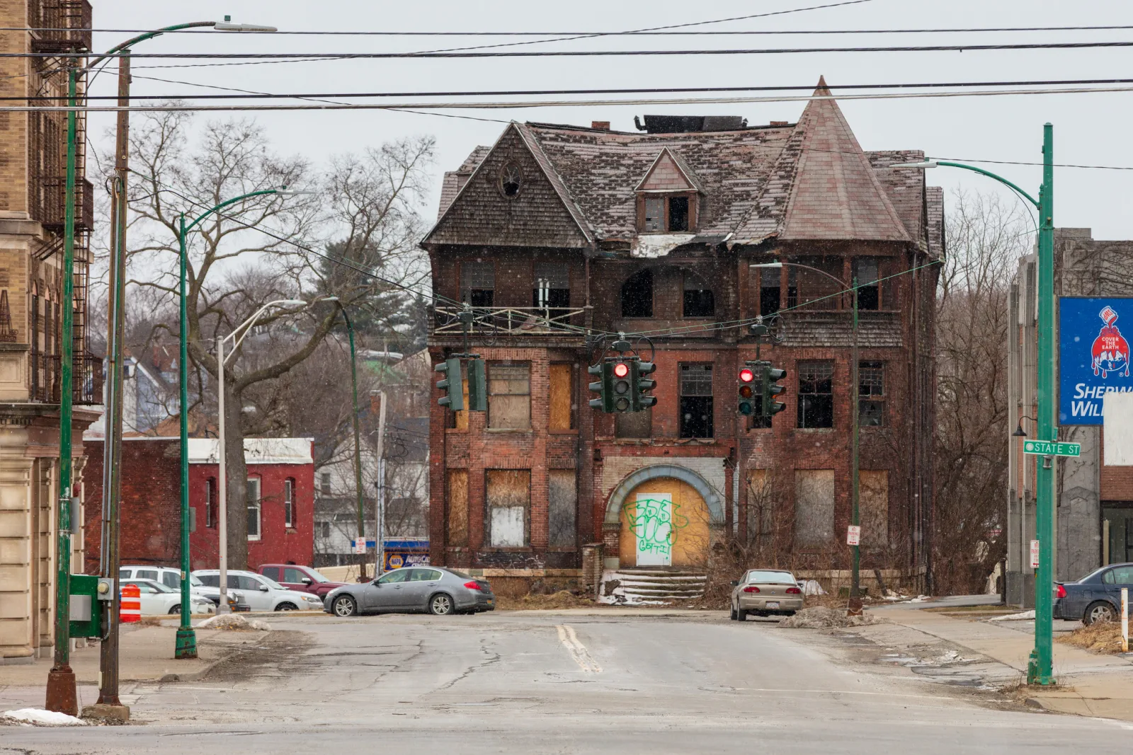 A derelict Victorian brick house with broken windows and a graffiti-covered boarded door at the end of a wide street, framed by street lights, traffic signals and parked cars
