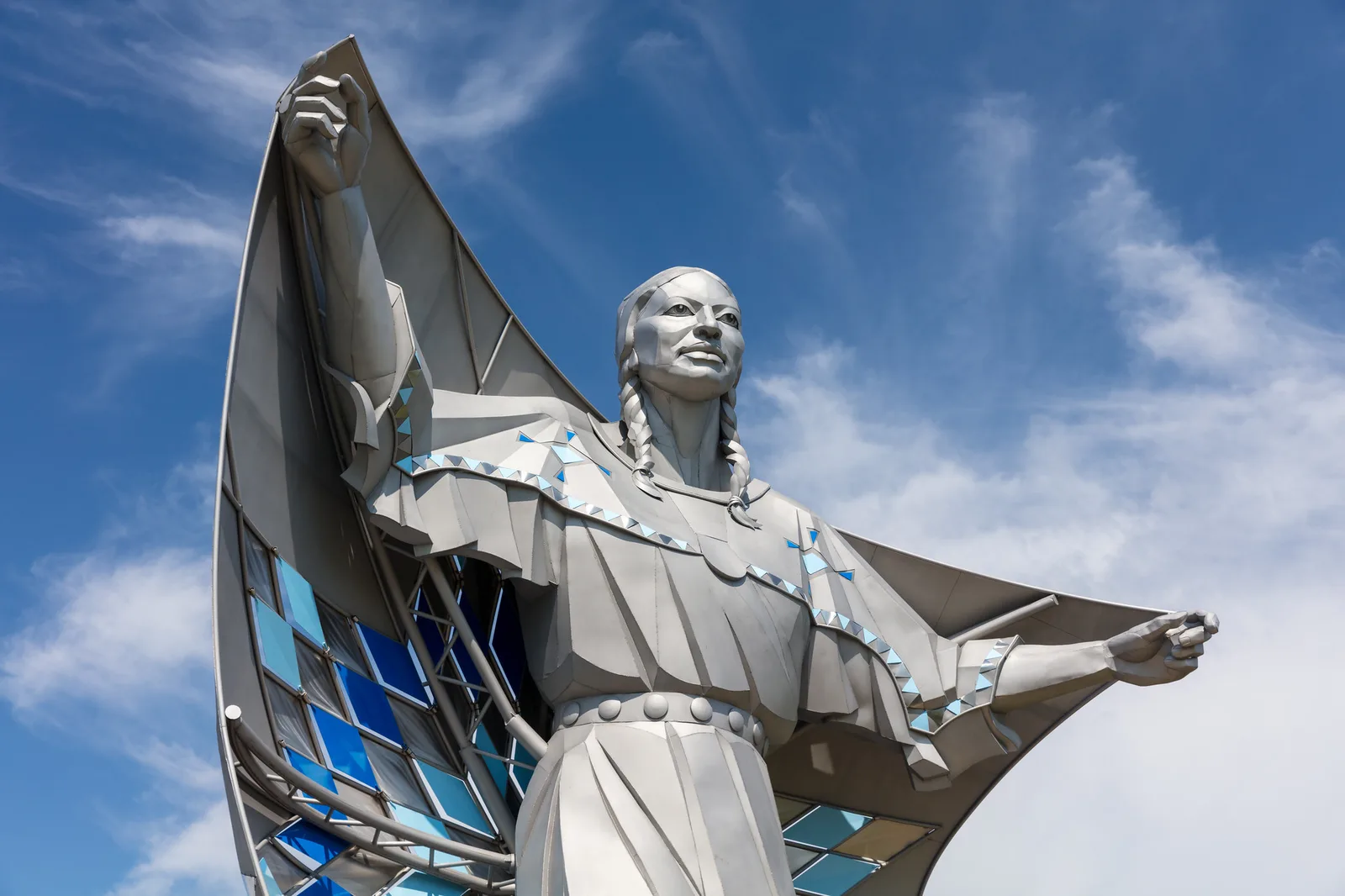 A tall stainless steel statue of a woman holding a star quilt open behind her against a blue sky, her face lit by high sun from the right with a small shadow beside the nose
