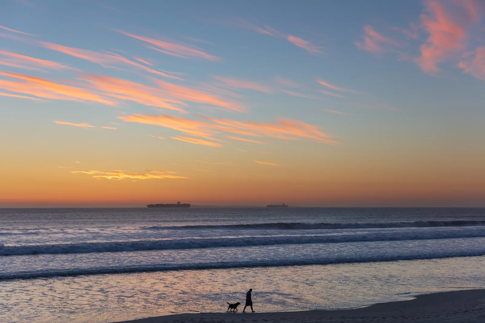 A person and a dog walking along the shoreline as small silhouettes at dusk, with rows of low waves, two ships on the horizon and pink clouds in an orange and blue sky