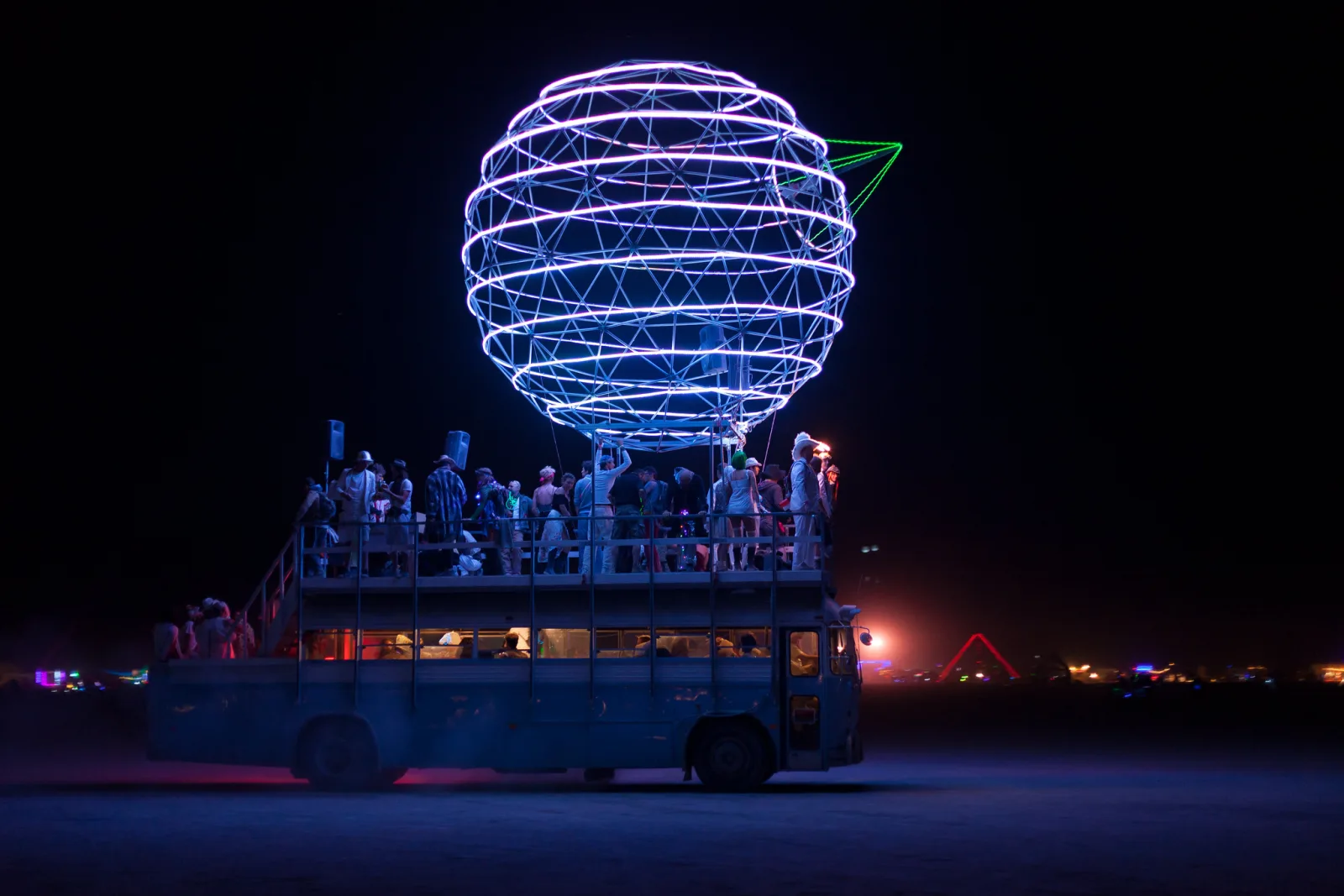 A tall vehicle carrying a large glowing white geodesic sphere, with people standing on its upper deck against a black night sky