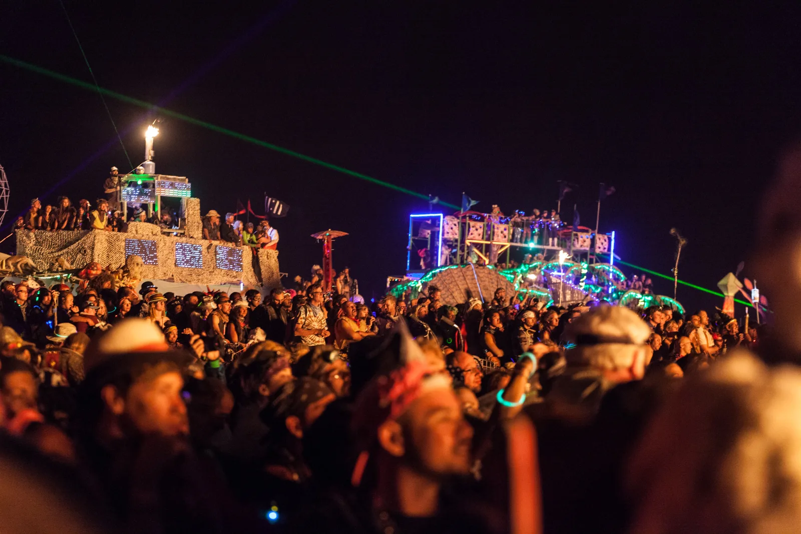 A large crowd at night lit by warm orange light, faces turned toward illuminated vehicles and structures in the background, with a green laser beam crossing the sky