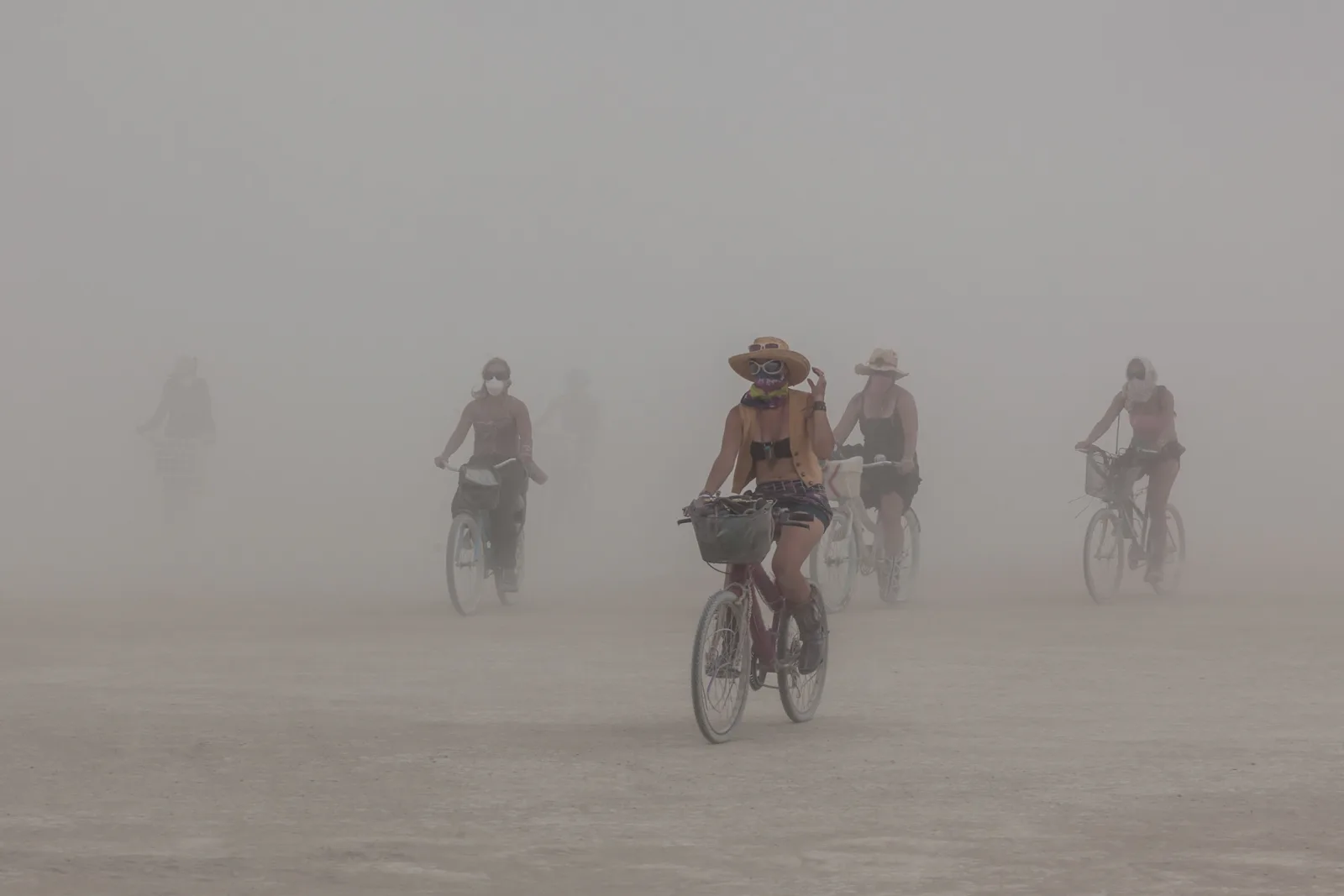 Cyclists in hats and dust masks riding toward the camera through a pale desert dust storm, the farthest riders fading into the haze