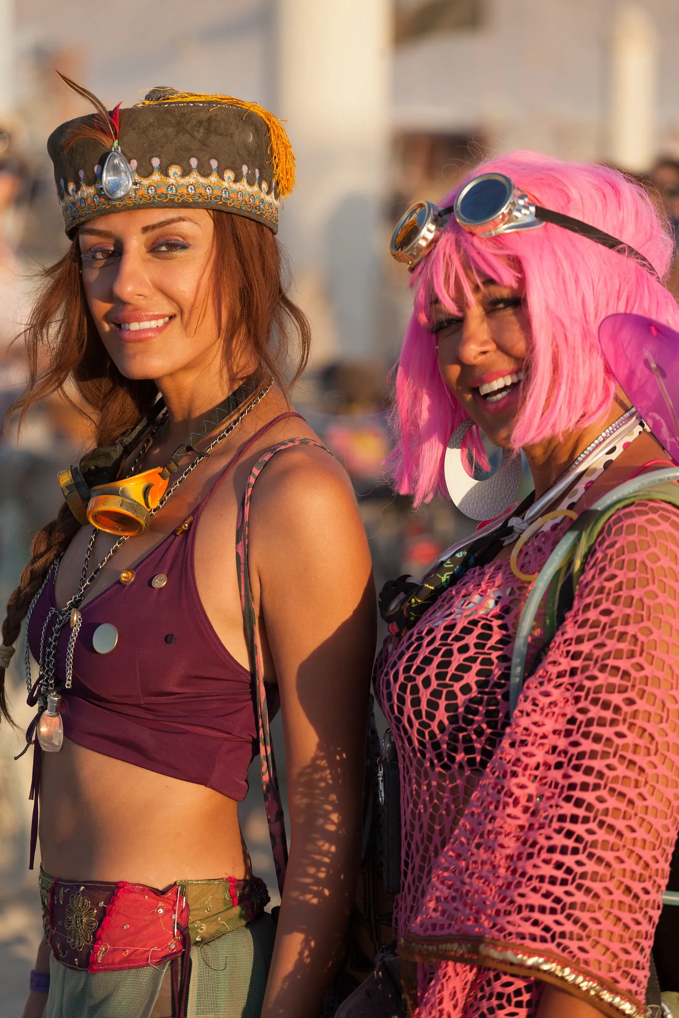 Two smiling women in festival costumes, one in a feathered cap and one in a pink wig and goggles, standing close together in warm low sunlight and looking into the camera