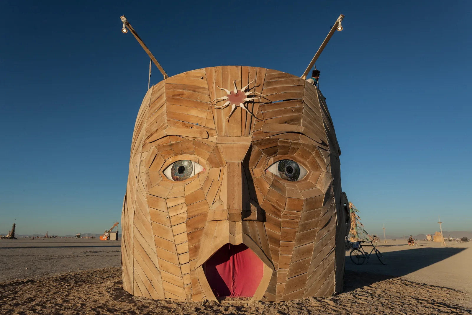 A large carved wooden face sculpture with antennae and a red tongue standing on flat desert ground under a deep blue sky