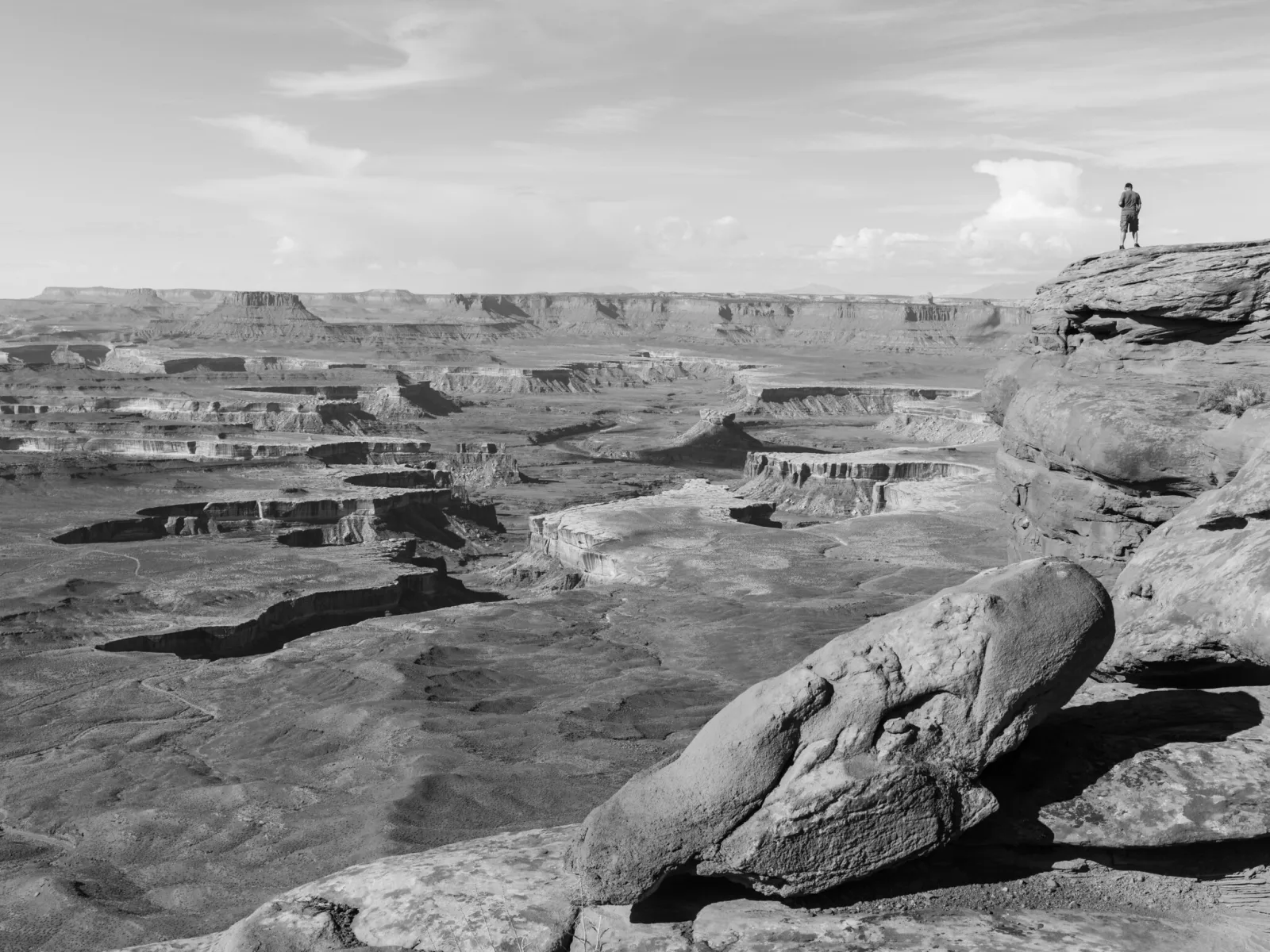 Black and white canyon landscape with a small figure standing on a rock ledge at upper right, silhouetted against the sky above the distant horizon