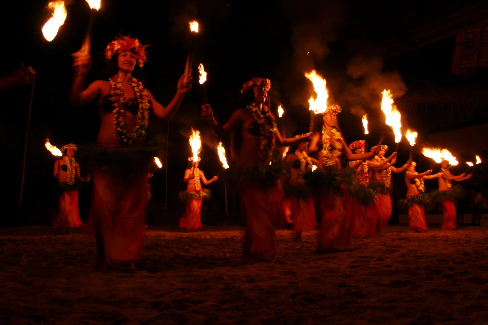A troupe of dancers in grass skirts perform at night holding flaming torches, their bodies lit orange by the fire against a dark background