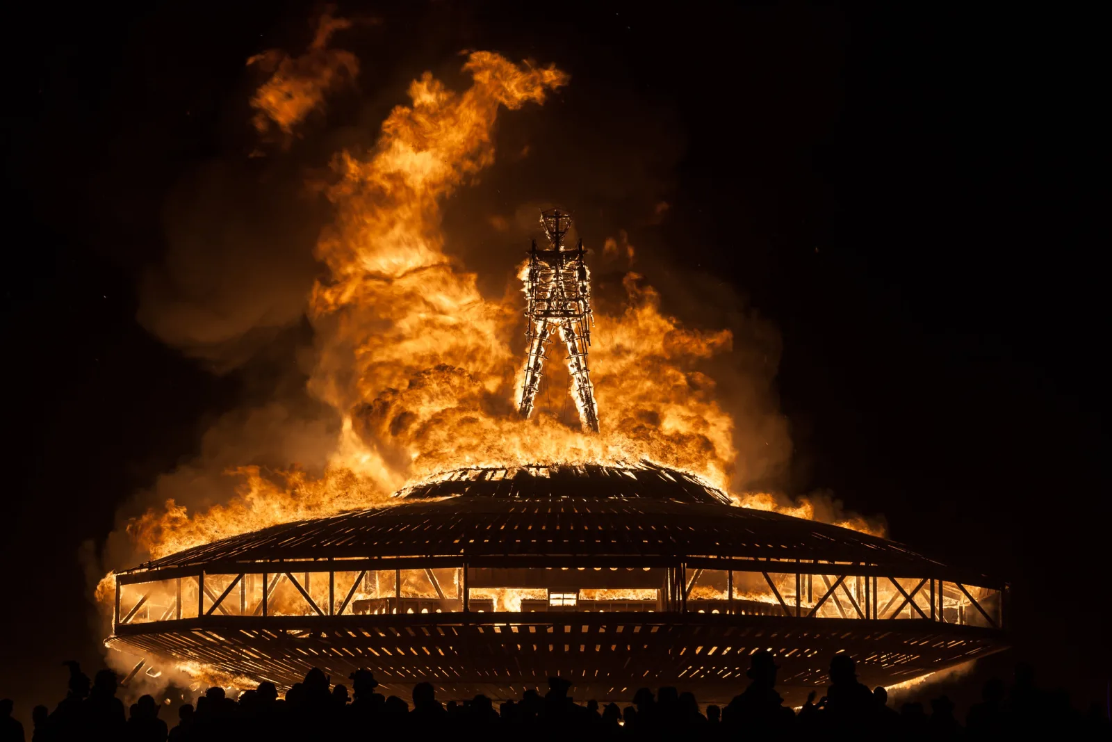A huge fire engulfing a tall wooden figure on top of a burning round structure at night, with smoke billowing and a row of spectators silhouetted along the bottom of the frame