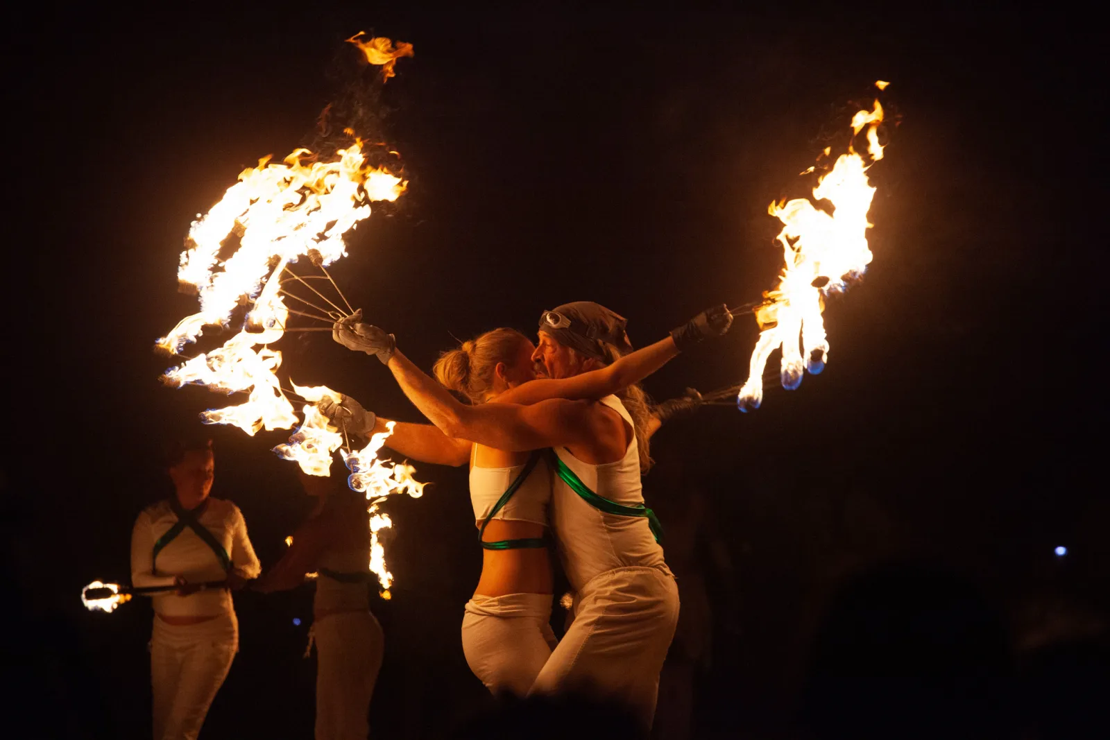 Two fire performers in white embracing between swinging flaming poi, with another performer holding a fire staff at the left edge, all against a black night background