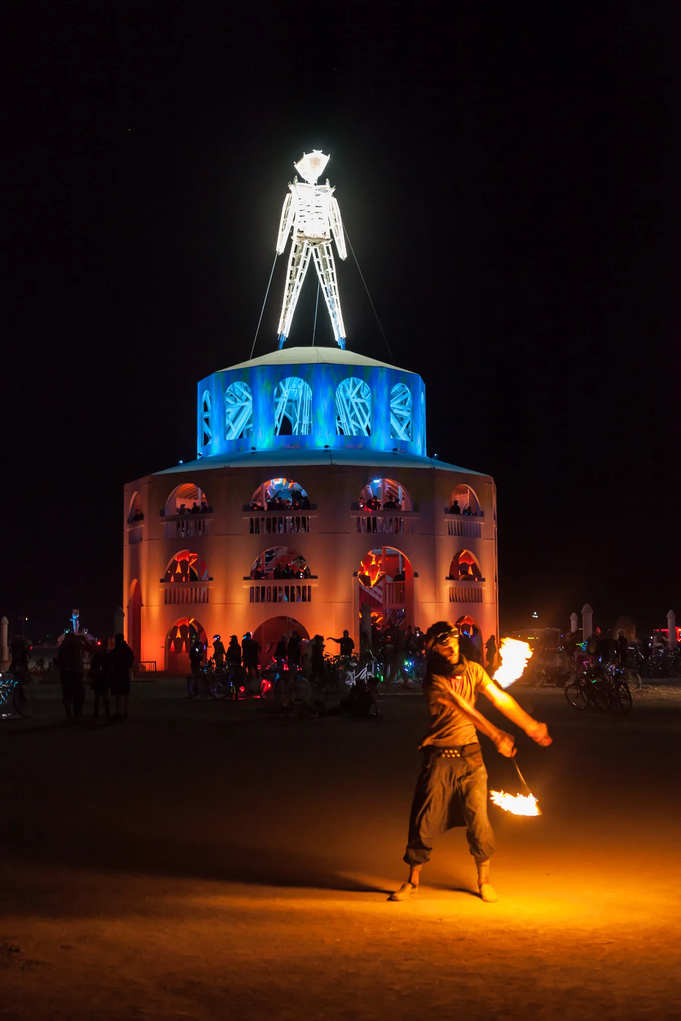 A fire spinner swinging flaming poi on open ground at night, with a lit tower topped by a glowing human figure behind, and the spinner's fire casting an orange pool of light on the ground