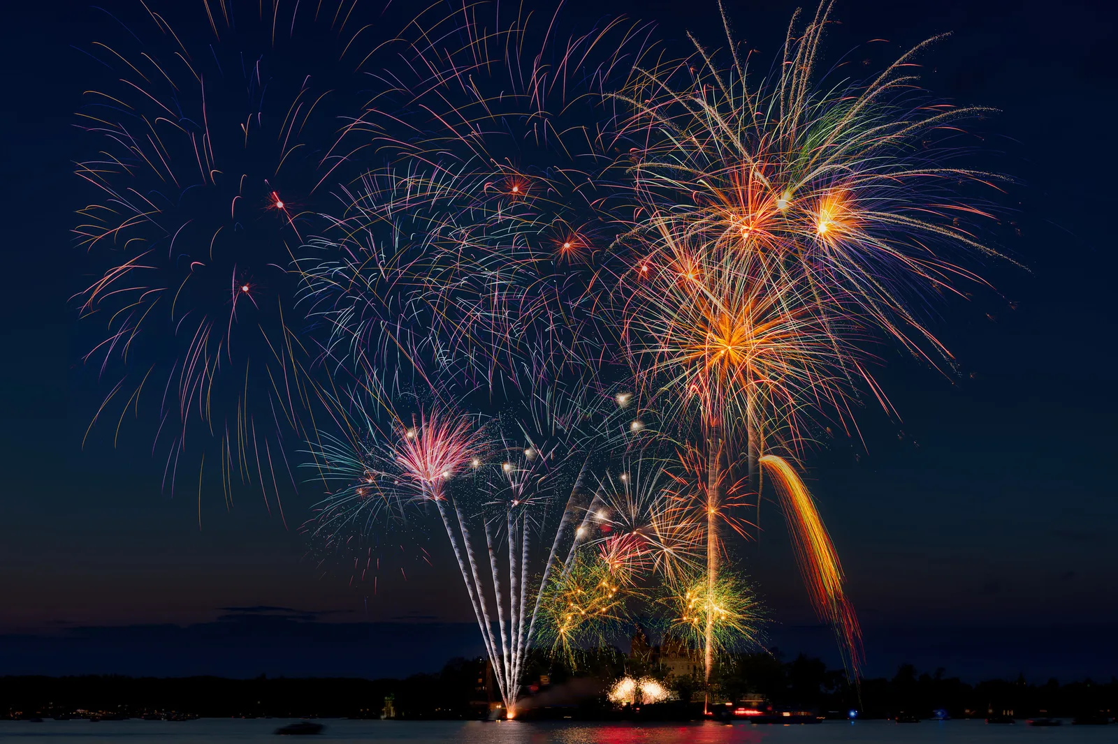 Several overlapping fireworks bursts in red, orange, green and white above a dark lake at night, with a thin band of twilight on the horizon