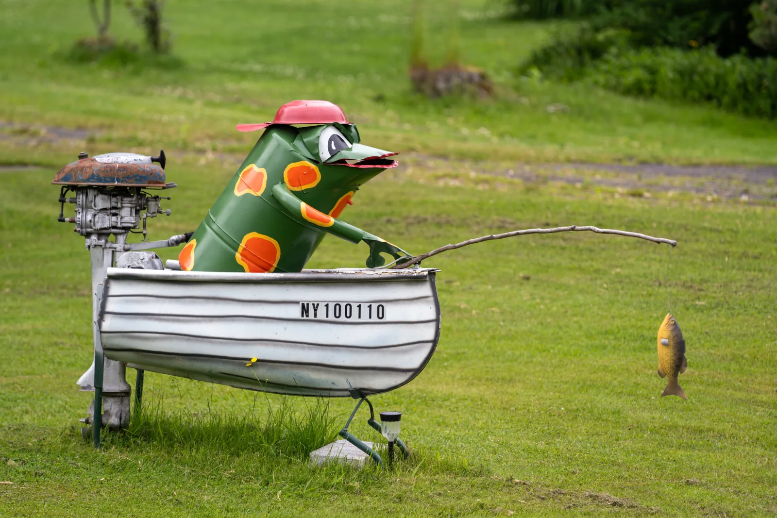 A painted green fish figure wearing a red cap sits in a small white boat on a lawn, holding a fishing rod with a fish hanging from the line