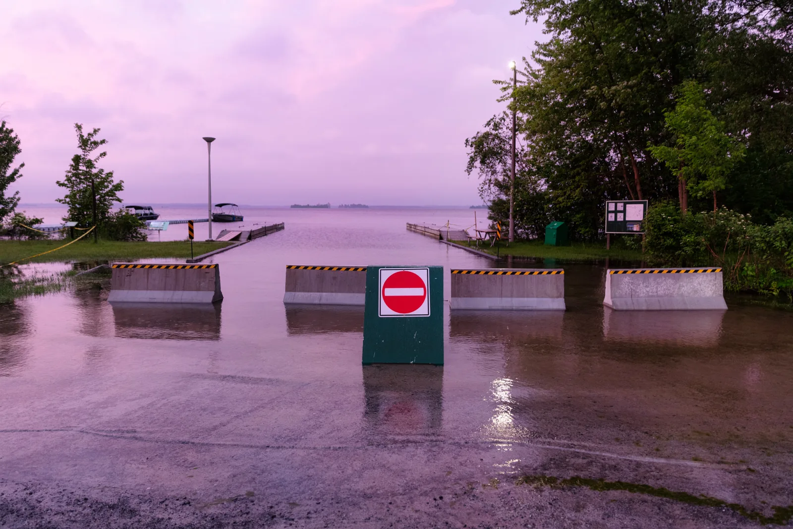 A lakeside parking lot under shallow floodwater at dusk, with a do not enter sign standing in the water and concrete barriers behind it