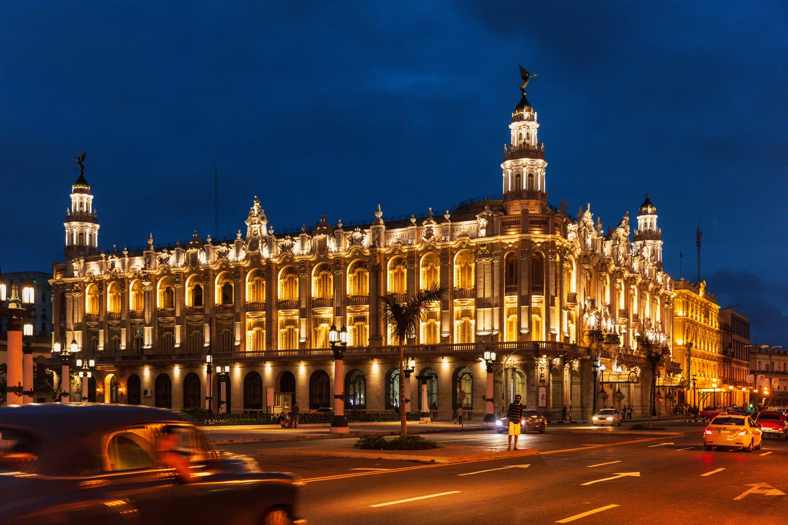 A large ornate floodlit theatre with towers and statues under a deep blue twilight sky, with blurred cars and light trails on the wide street in front