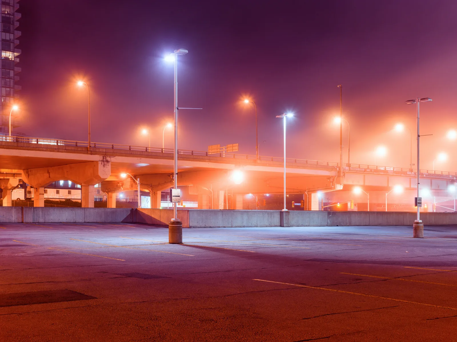 An empty parking lot under an overpass on a foggy night, lit by a mix of orange and bluish-white street lights