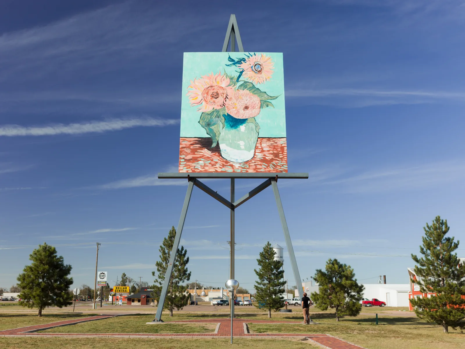 An enormous reproduction of a sunflower painting on a giant steel easel beside a road, with a single person standing at its base